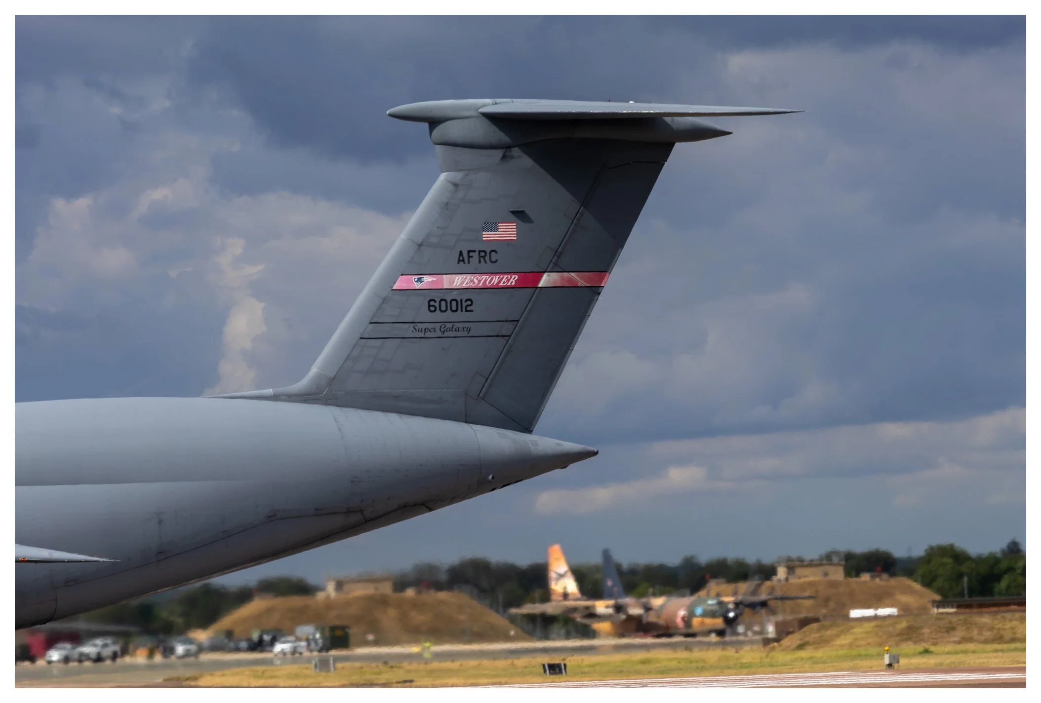 Close-up view of the tail of a military aircraft with markings, parked on a runway with a cloudy sky overhead and other aircraft and vehicles in the background.