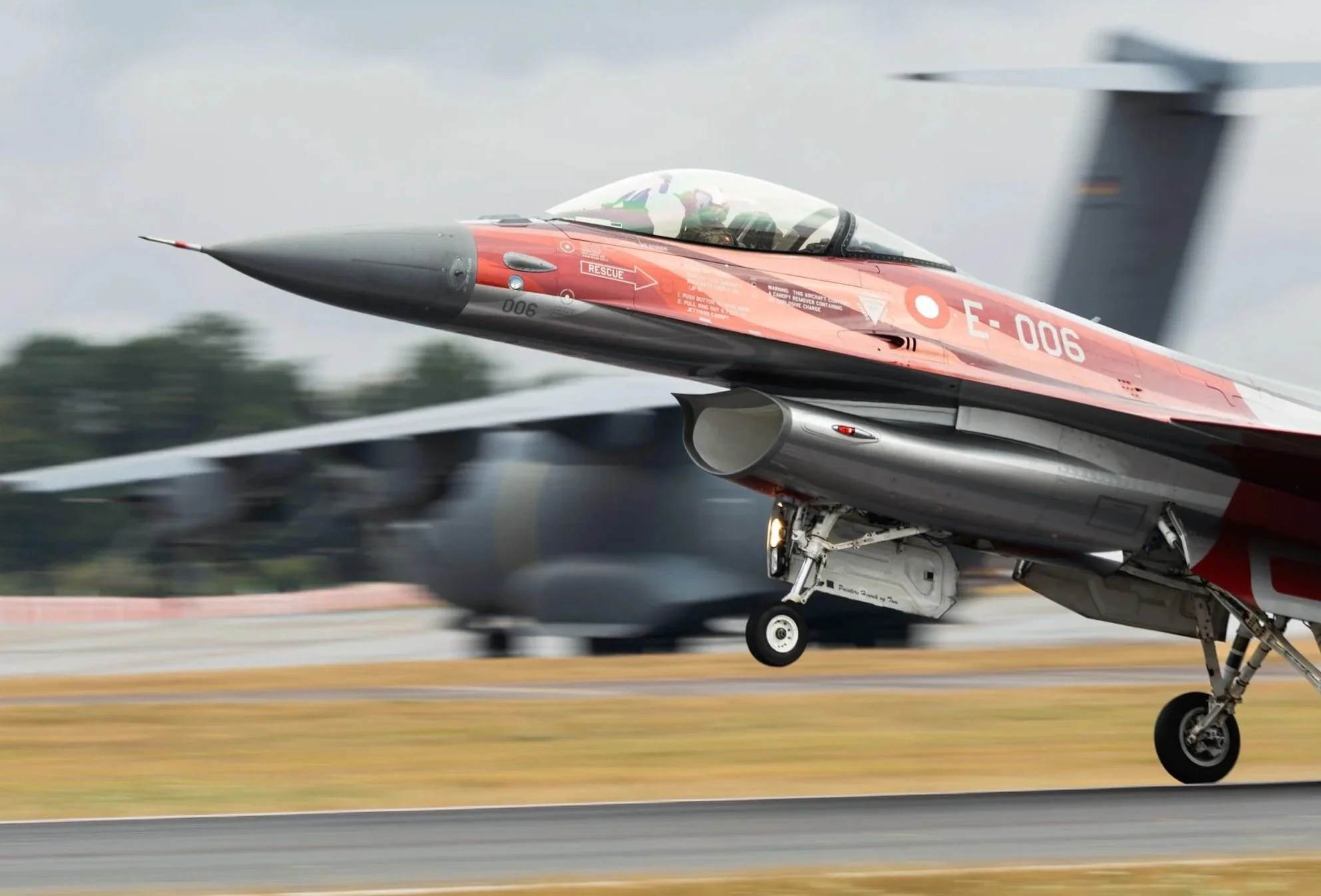 A military fighter jet aircraft taking off from a runway with blurred background of another jet and trees.