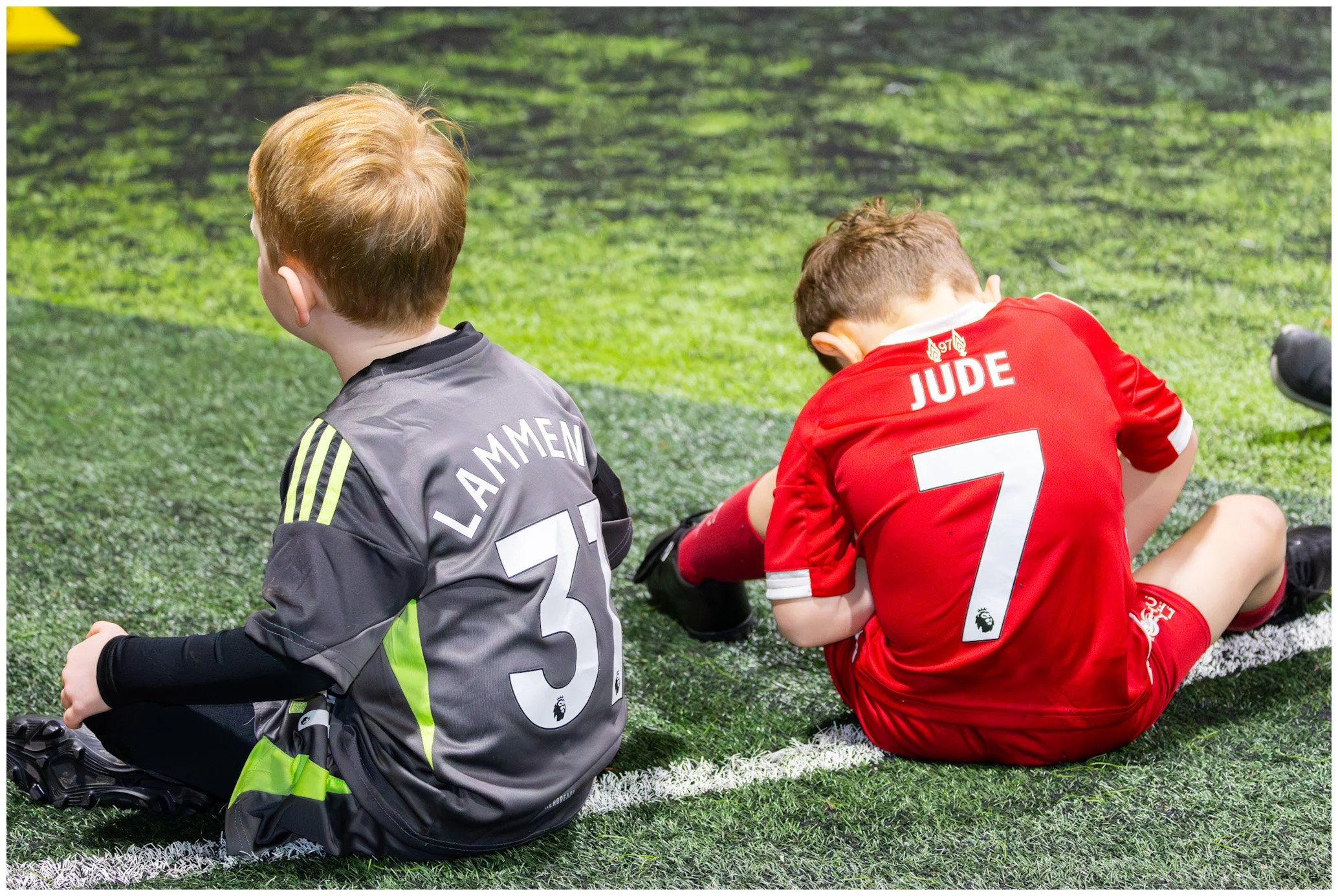 Two young boys sitting on a green soccer field; one wearing a gray jersey with 'LAMMEN' and number 31, the other wearing a red jersey with 'JUDE' and number 7.