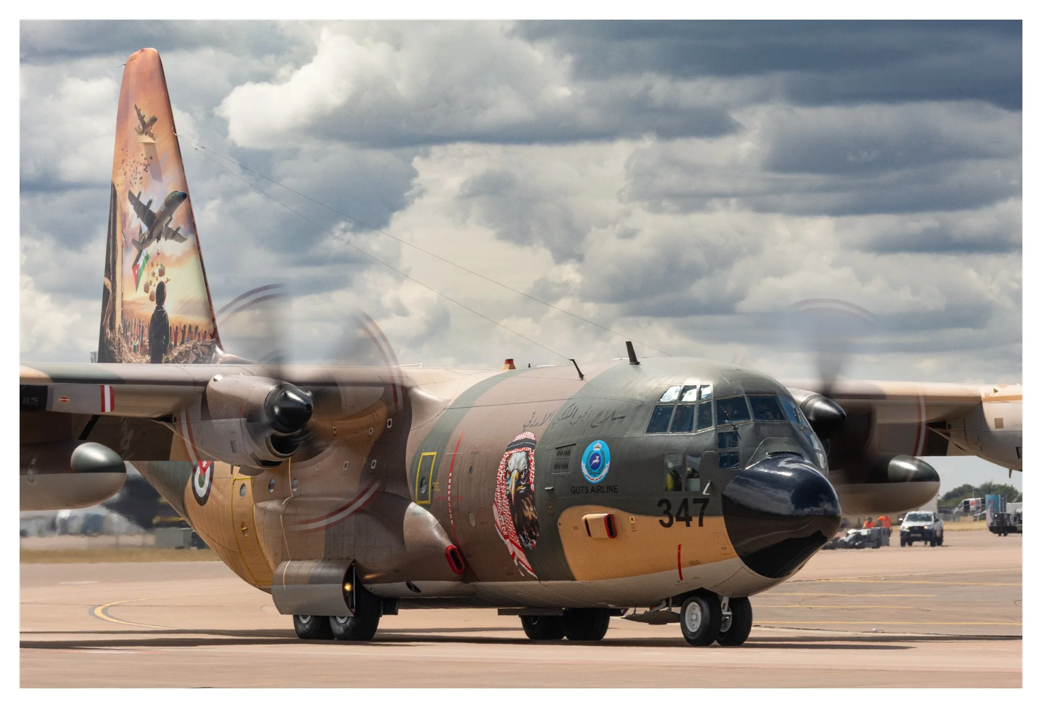 A military cargo aircraft on the tarmac with colorful artwork on its tail. The artwork depicts jets and a person watching the planes in a sky filled with clouds.