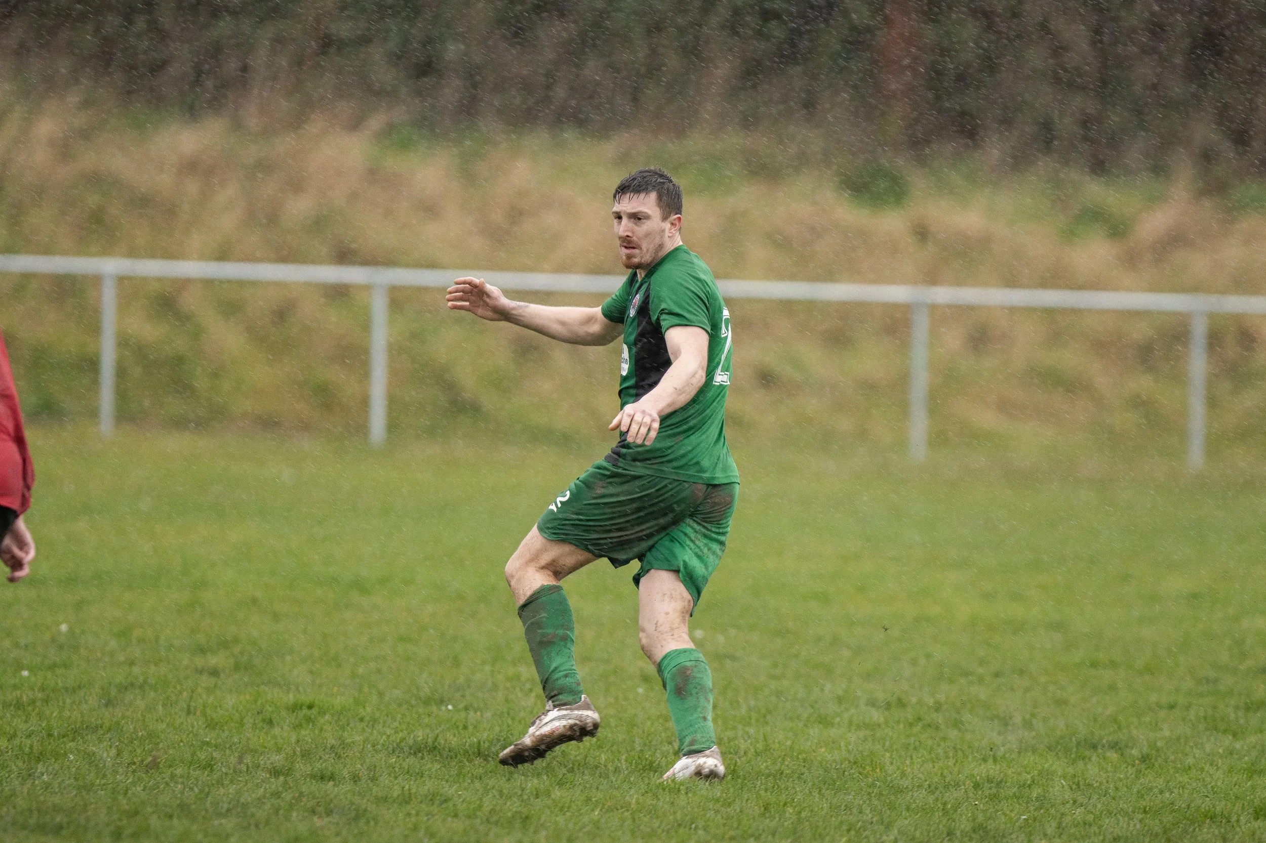 A male soccer player is on a grass field during rain, wearing a green jersey and shorts, with muddy cleats, in a defensive stance.