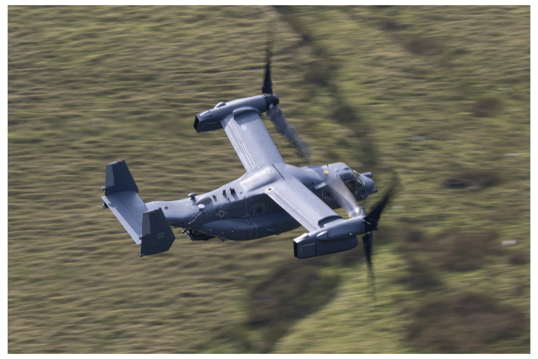 A military V-22 Osprey tiltrotor aircraft flying over a grassy landscape during daylight.