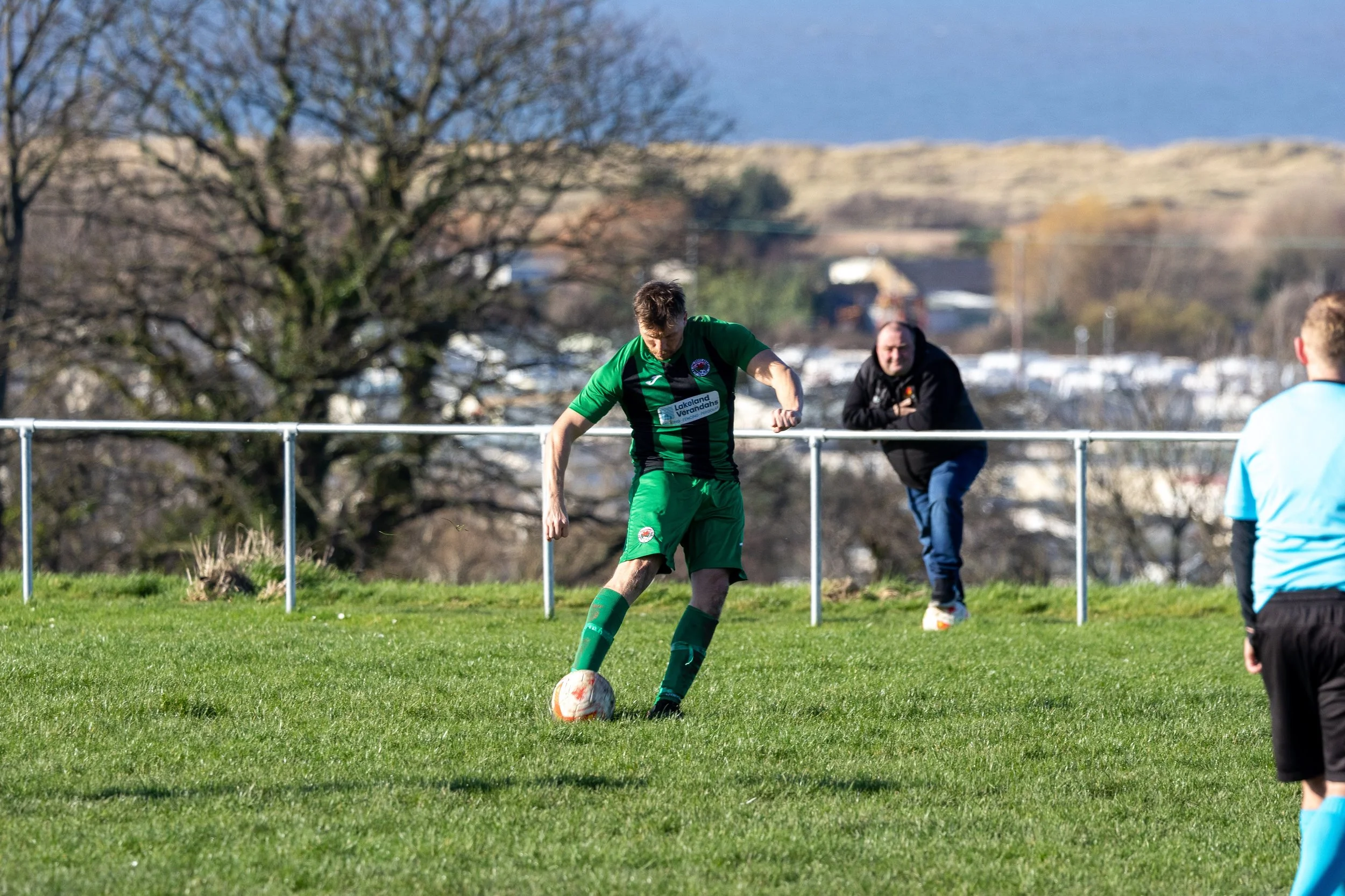 A soccer player in a green and black kit kicking a soccer ball on a grassy field during a game, with a man in a black hoodie watching from behind a white fence in the background, and another player in a light blue jersey in the foreground.