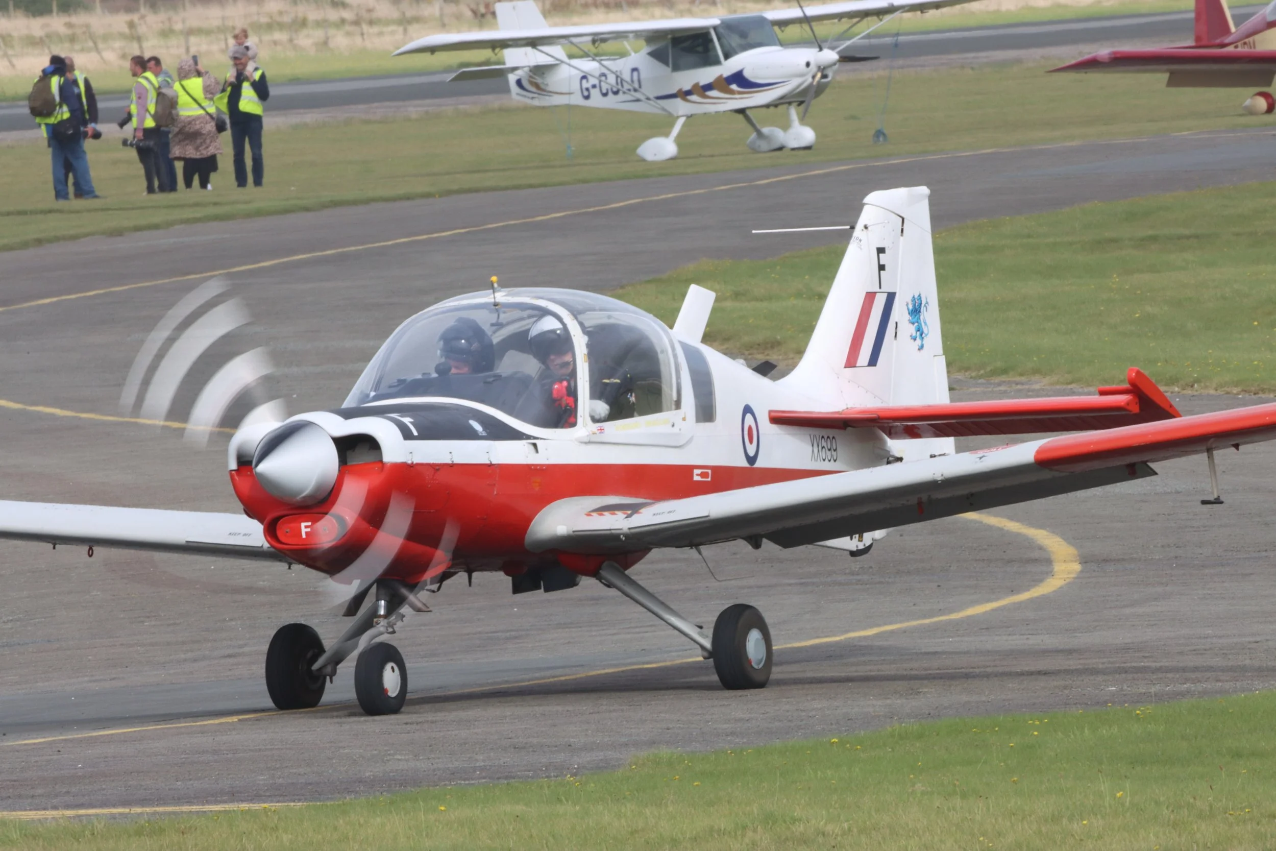 A small propeller aircraft painted in red, white, and black, with a rounded nose, landing on a runway. There is a second plane in the background with people standing nearby on the grass.