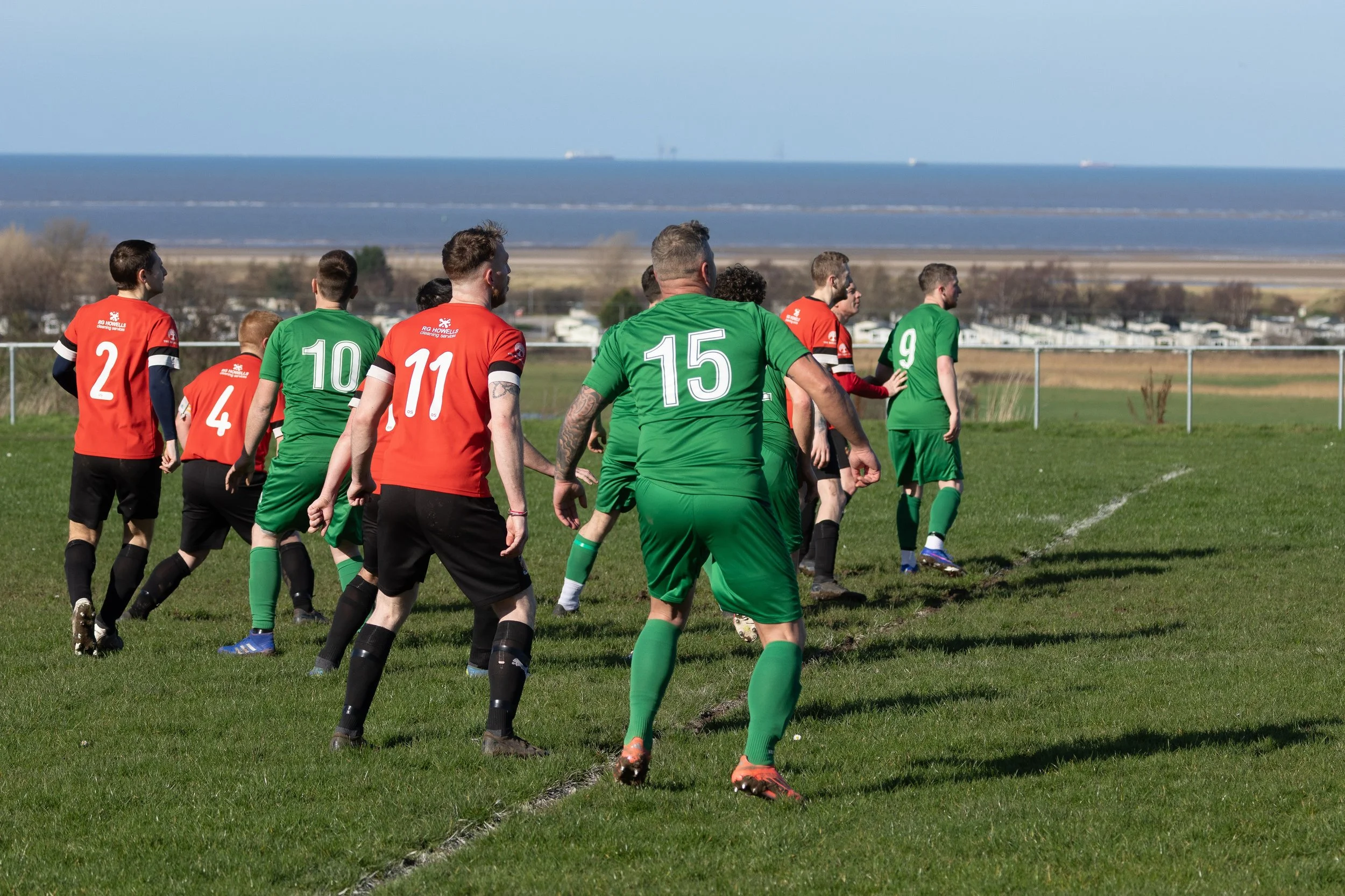 Soccer match with players in red and green uniforms on a grassy field near a beach, with ocean and boats in the distance.