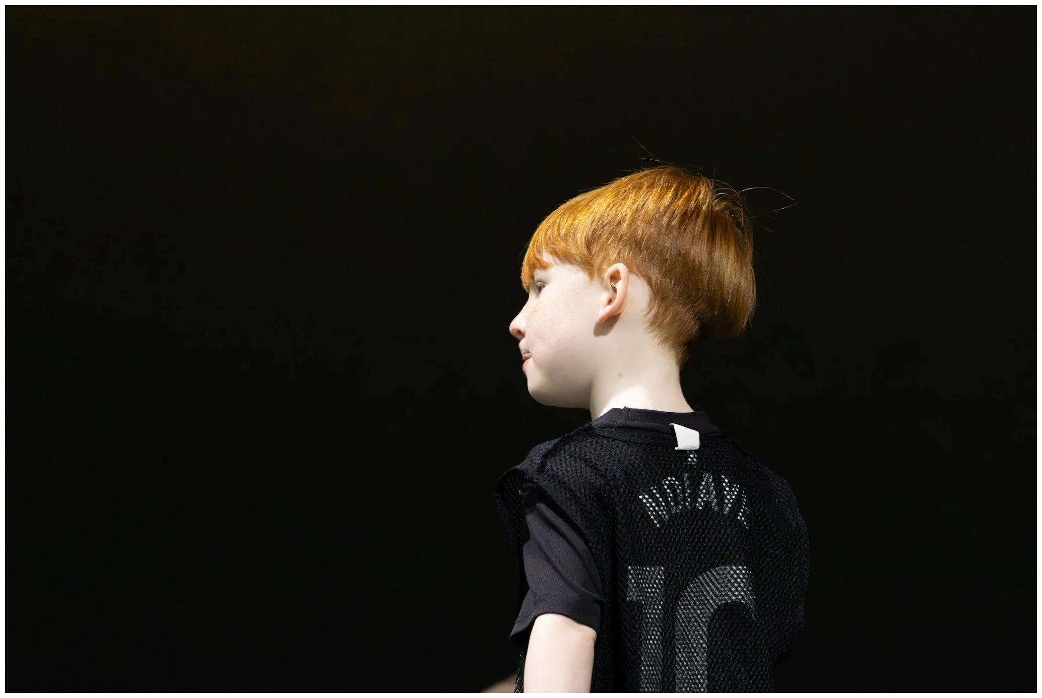 Side view of a young boy with red hair wearing a black sports jersey with the number 10 on the back, standing on a dark background.