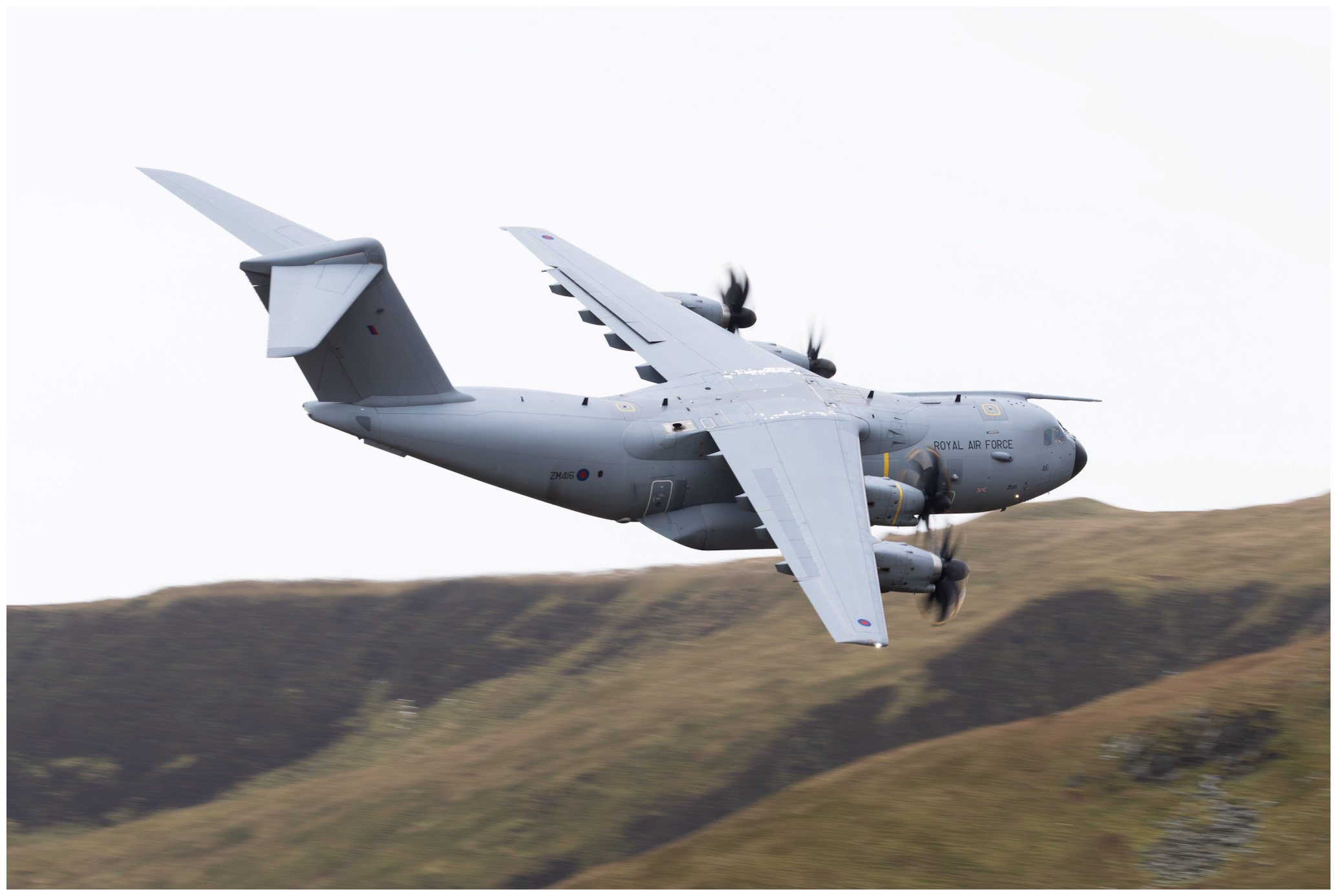A gray Royal Air Force military transport aircraft flying low over a grassy hillside.