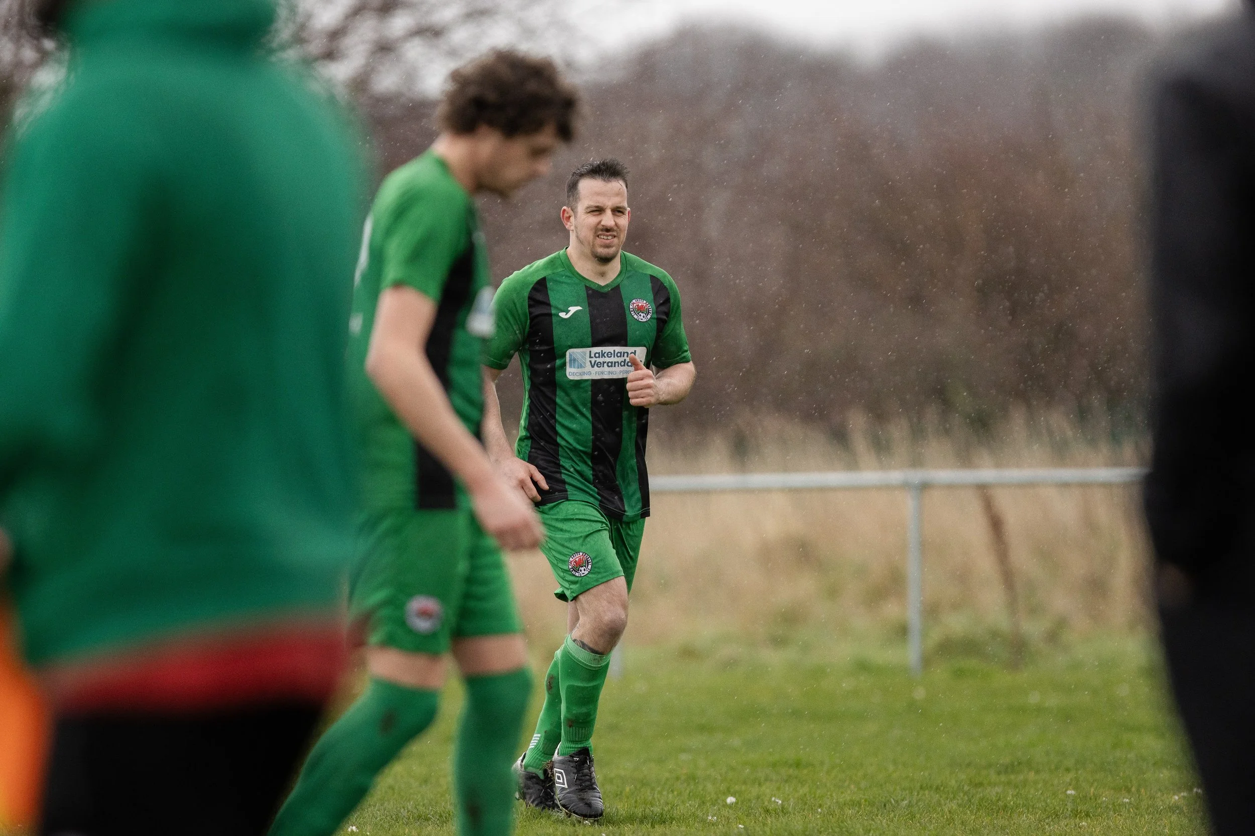 A group of soccer players in green and black uniforms on the field, with one player in focus in the background, running or jogging.