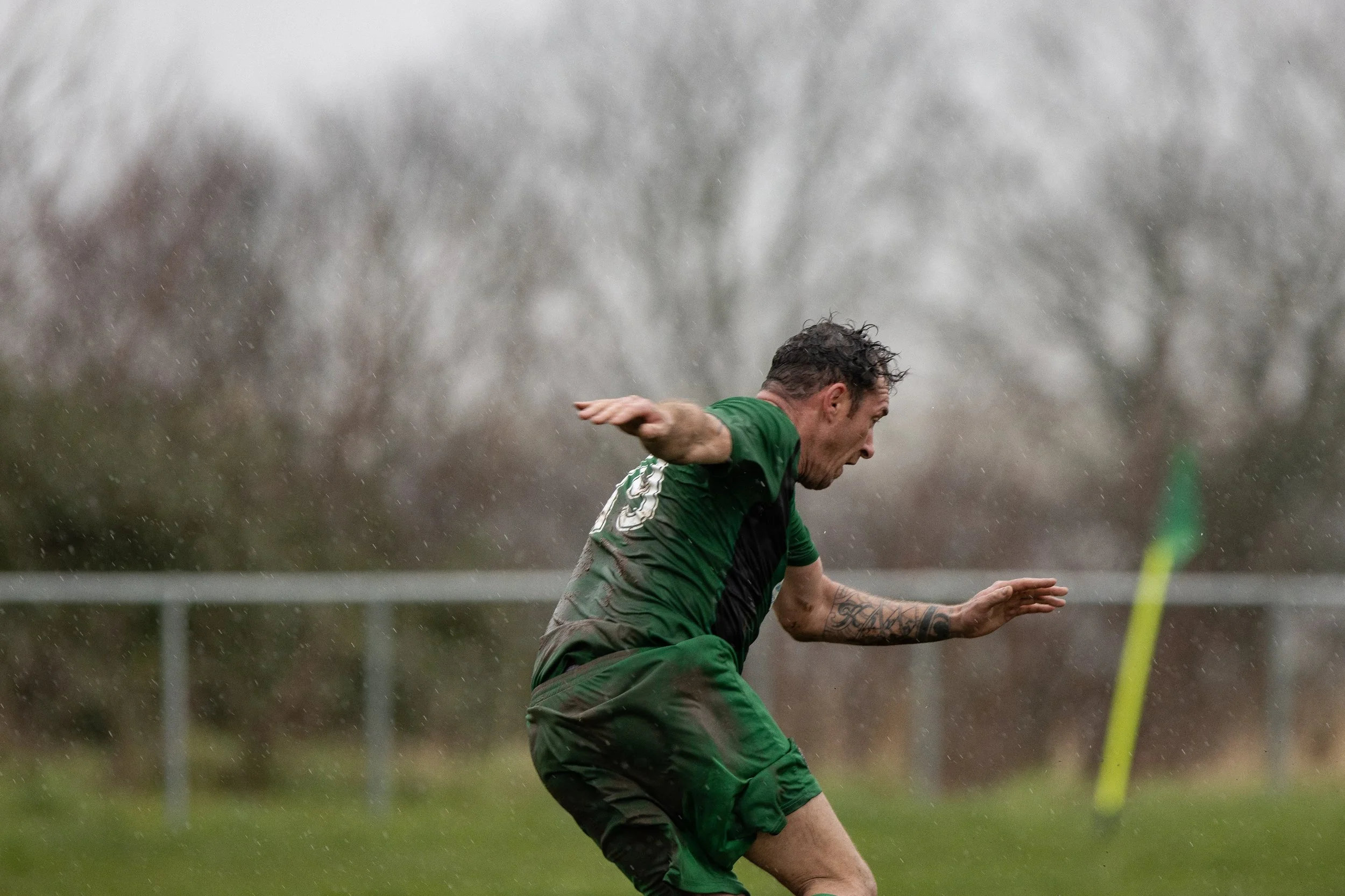 A soccer player wearing a green and black uniform is playing on a muddy field in rainy weather.