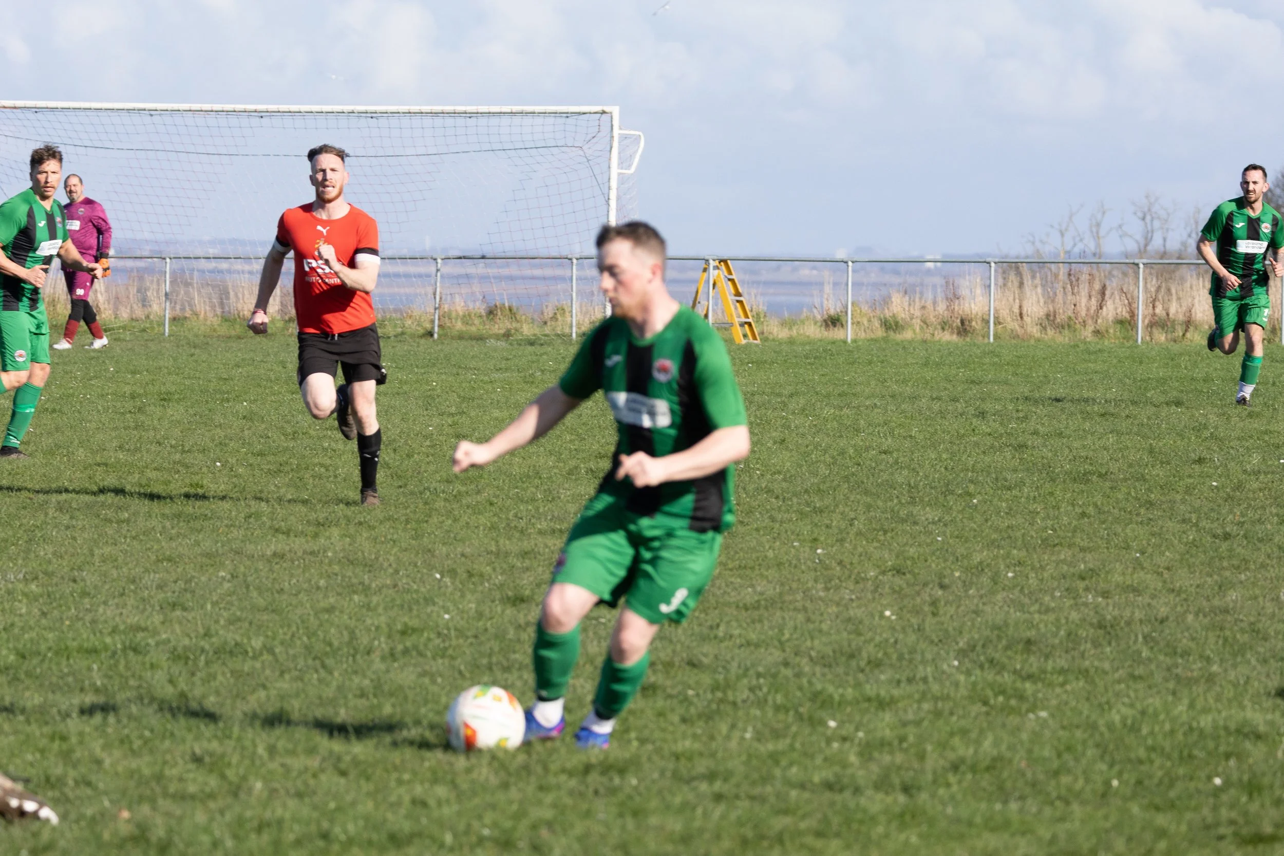 Soccer players on a green field, one in the foreground about to kick a soccer ball, others running and standing in the background near a goalpost, with a cloudy sky and some trees behind the field.
