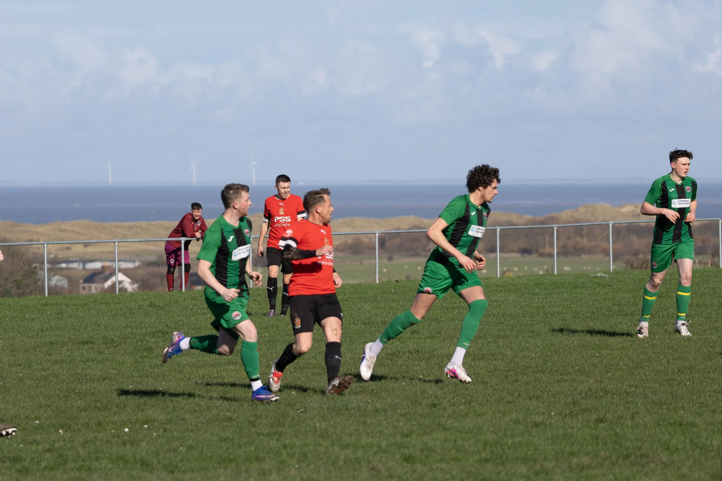 Soccer match with players in green and red uniforms on a grassy field during daytime, with ocean and wind turbines in the background.