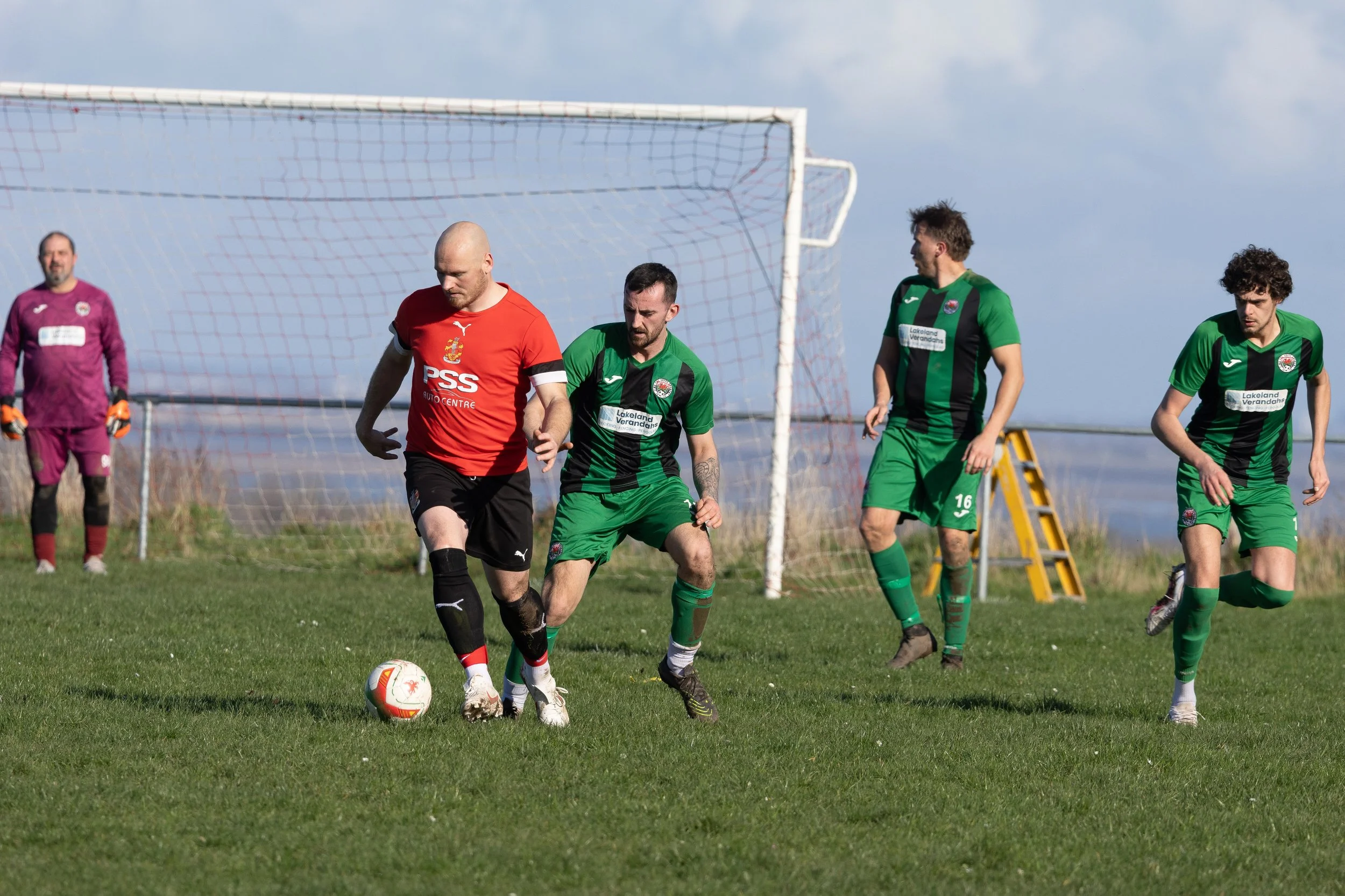 Four soccer players on a field near the goal, with one player in a red jersey controlling the ball, and three players in green jerseys approaching. A goalkeeper stands in front of the goal in a purple uniform.