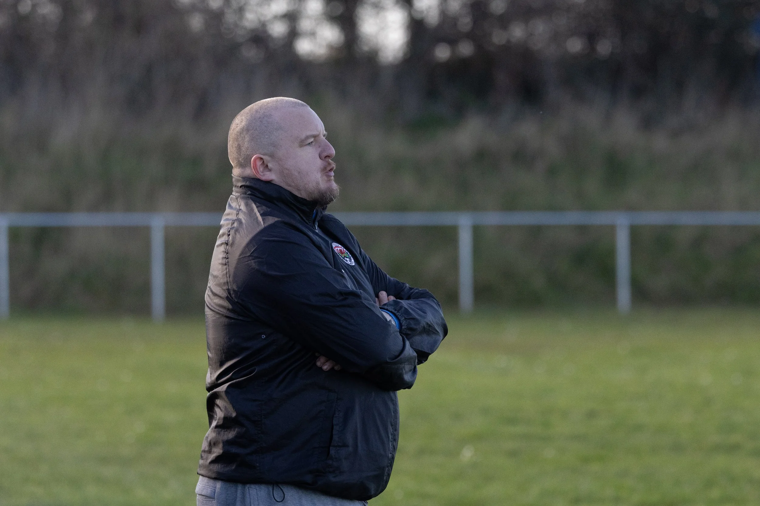 A man standing with arms crossed on a sports field, wearing a black jacket with a logo, and looking to the right, during daylight.