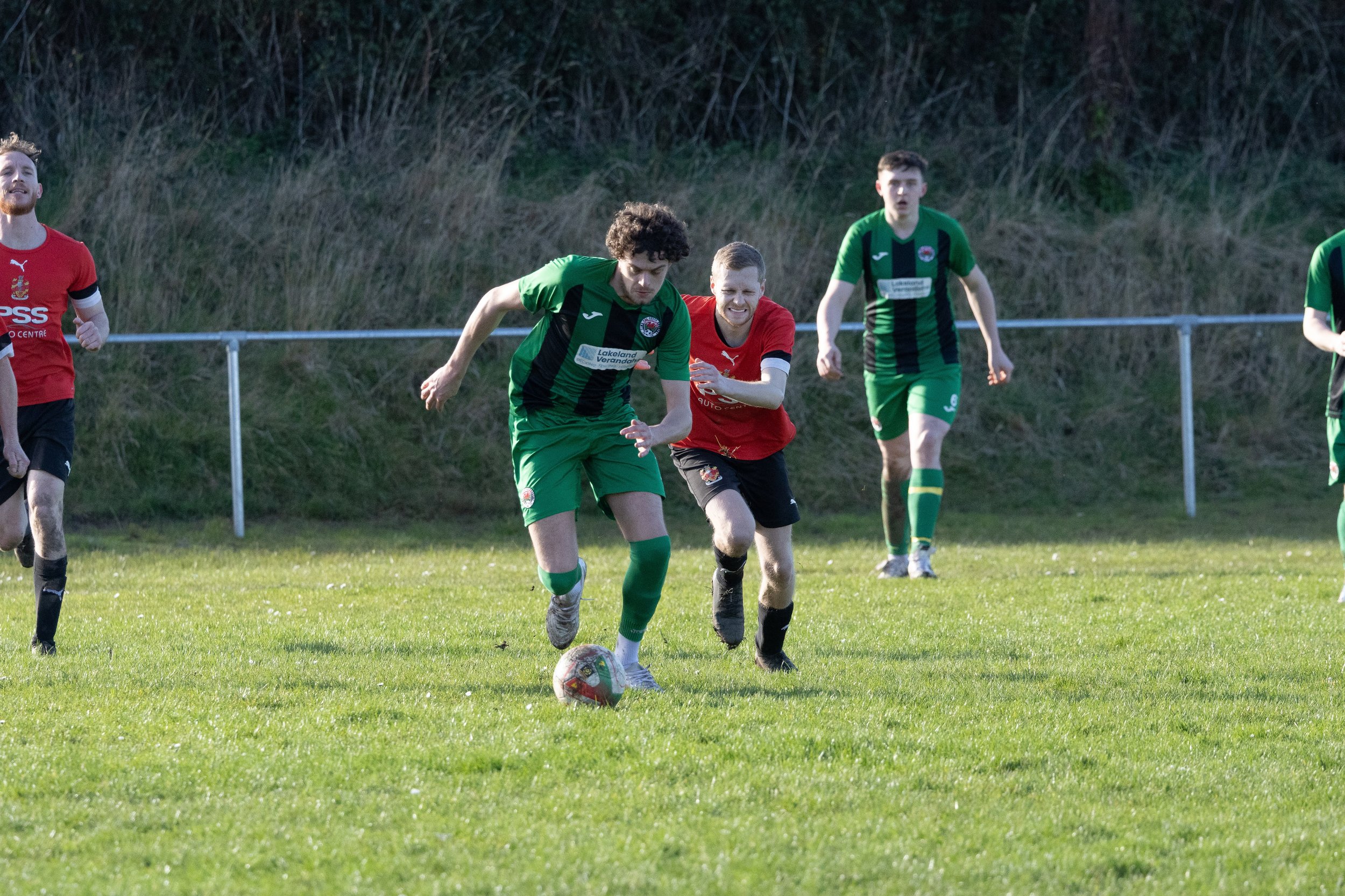 Soccer players in green and red jerseys competing for the ball on a grassy field with a hillside and trees in the background.
