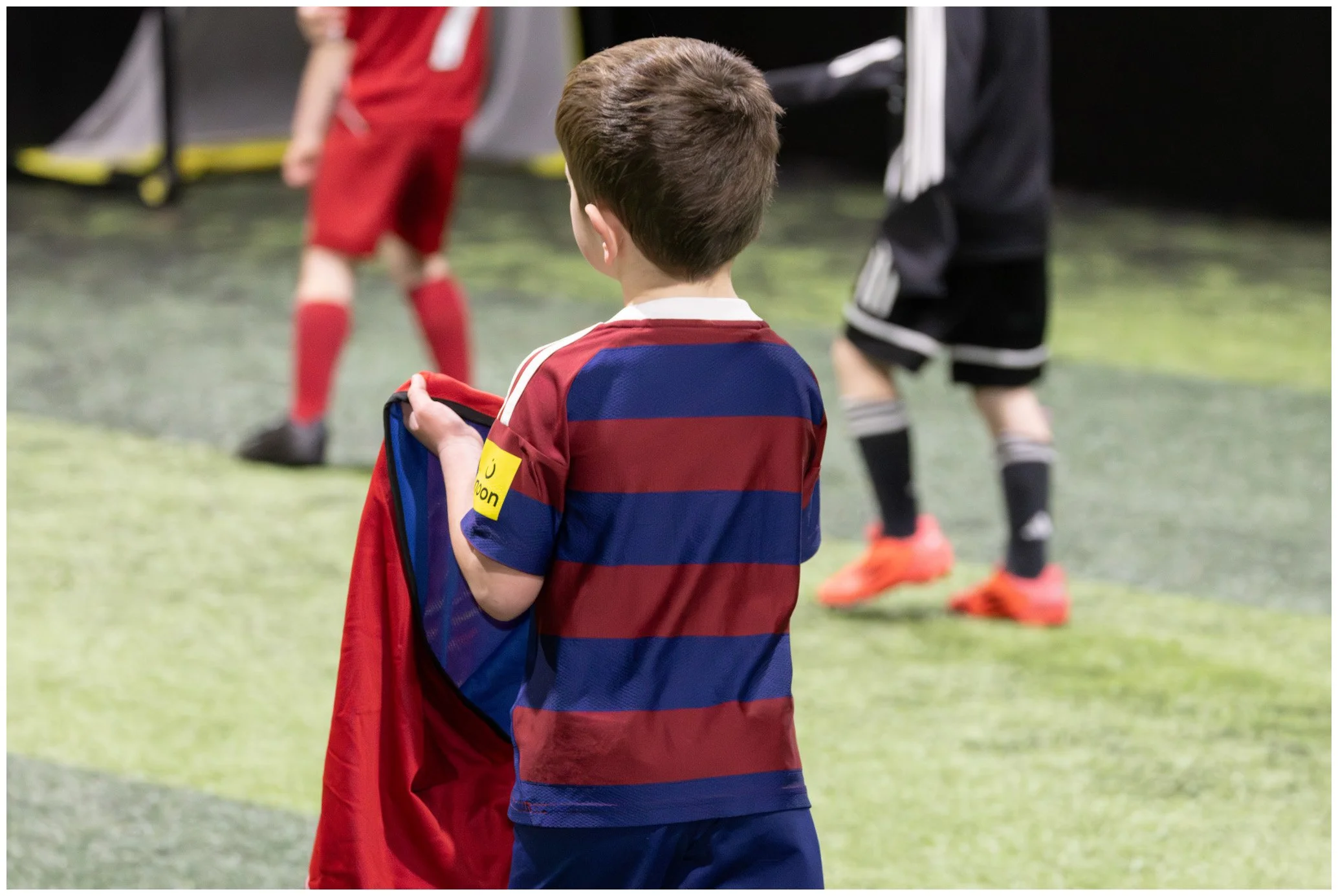 A young boy in a soccer uniform holding a red jersey, standing on an indoor soccer field while two other children in sportswear are nearby.