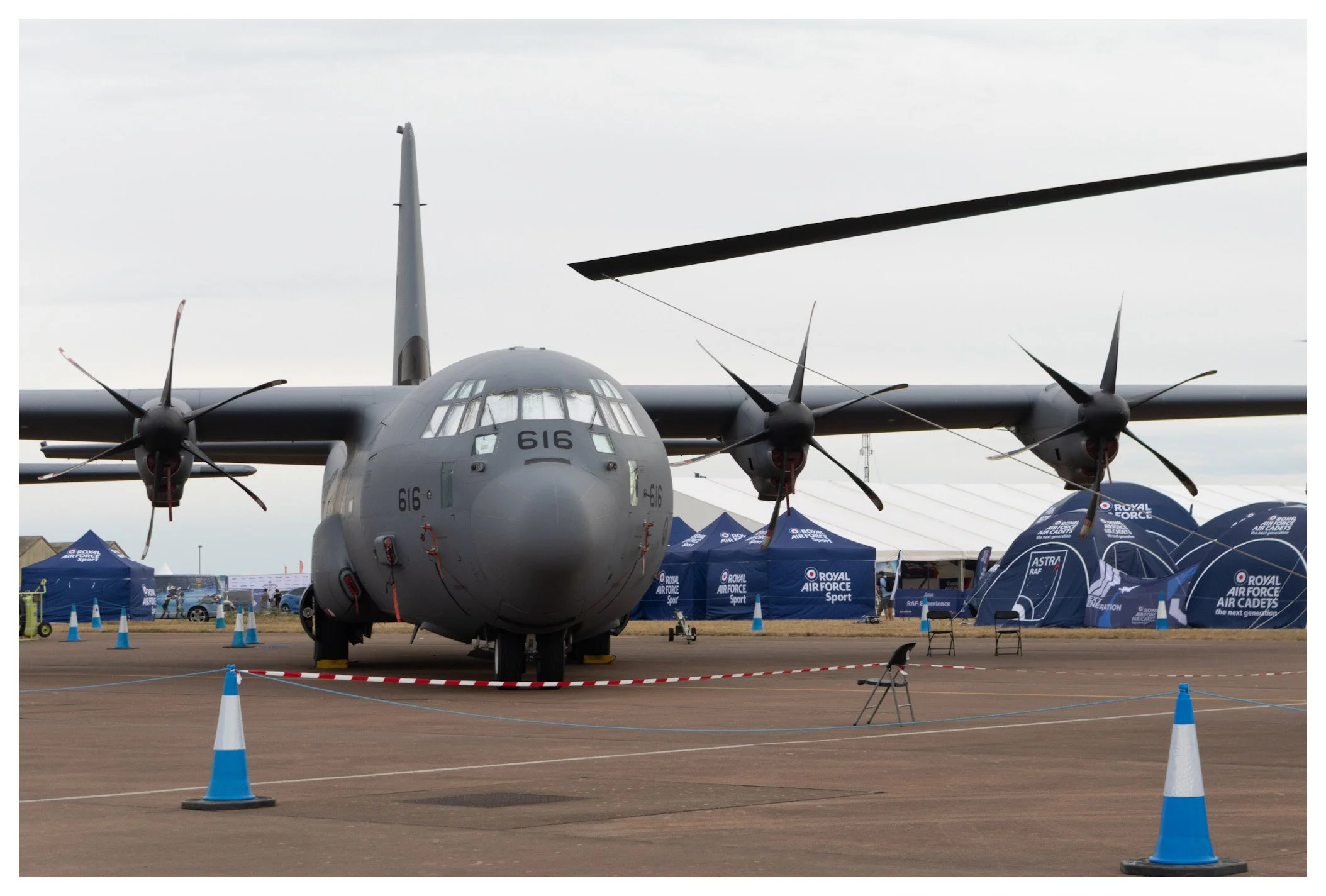 A military aircraft on display at an air show, surrounded by blue tents and orange traffic cones with a cloudy sky in the background.