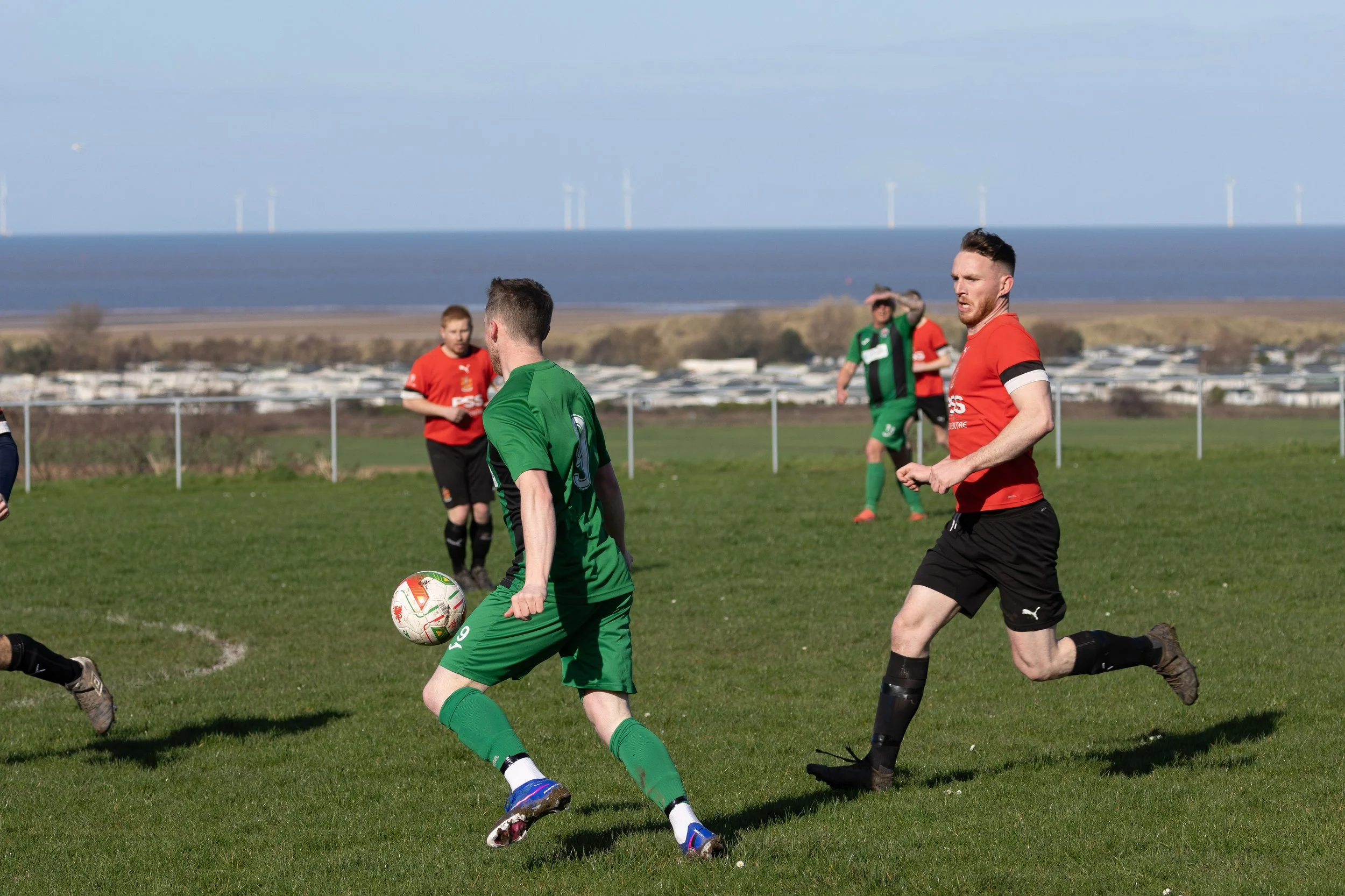 Soccer players in green and red jerseys playing on a grassy field near water and wind turbines.