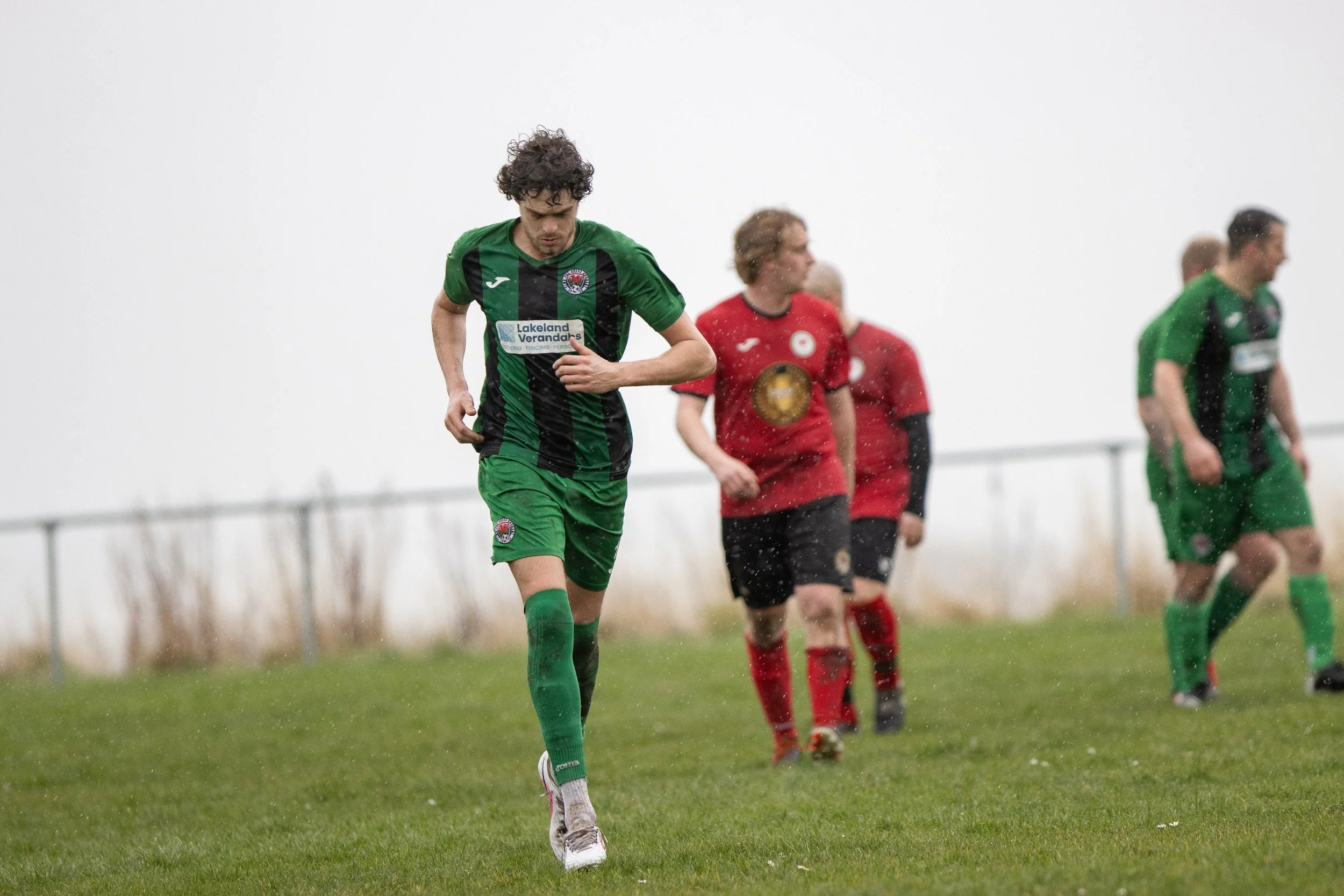 Soccer players walking on a rainy field, with one player in a green and black striped jersey in the foreground and others in red jerseys in the background.