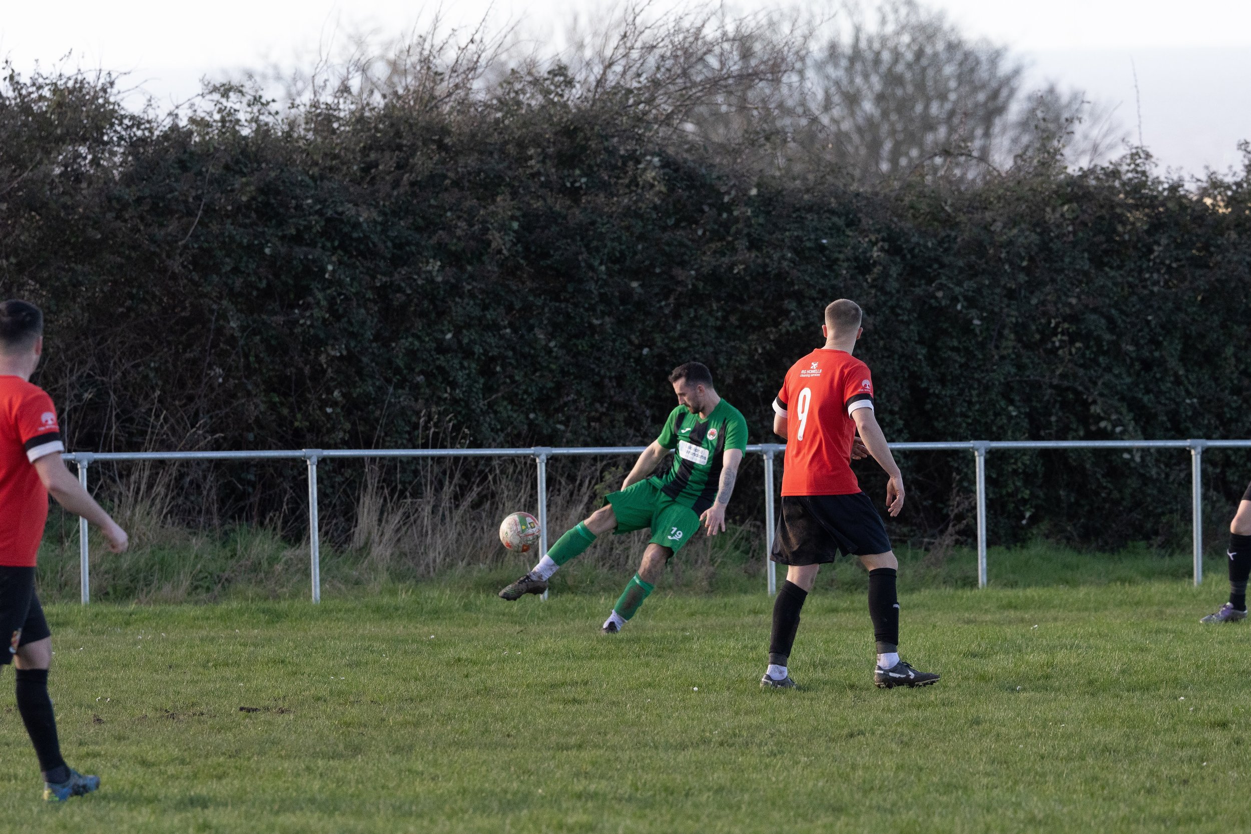 Soccer players in red and green uniforms on a grassy field with a black bush and trees in the background.