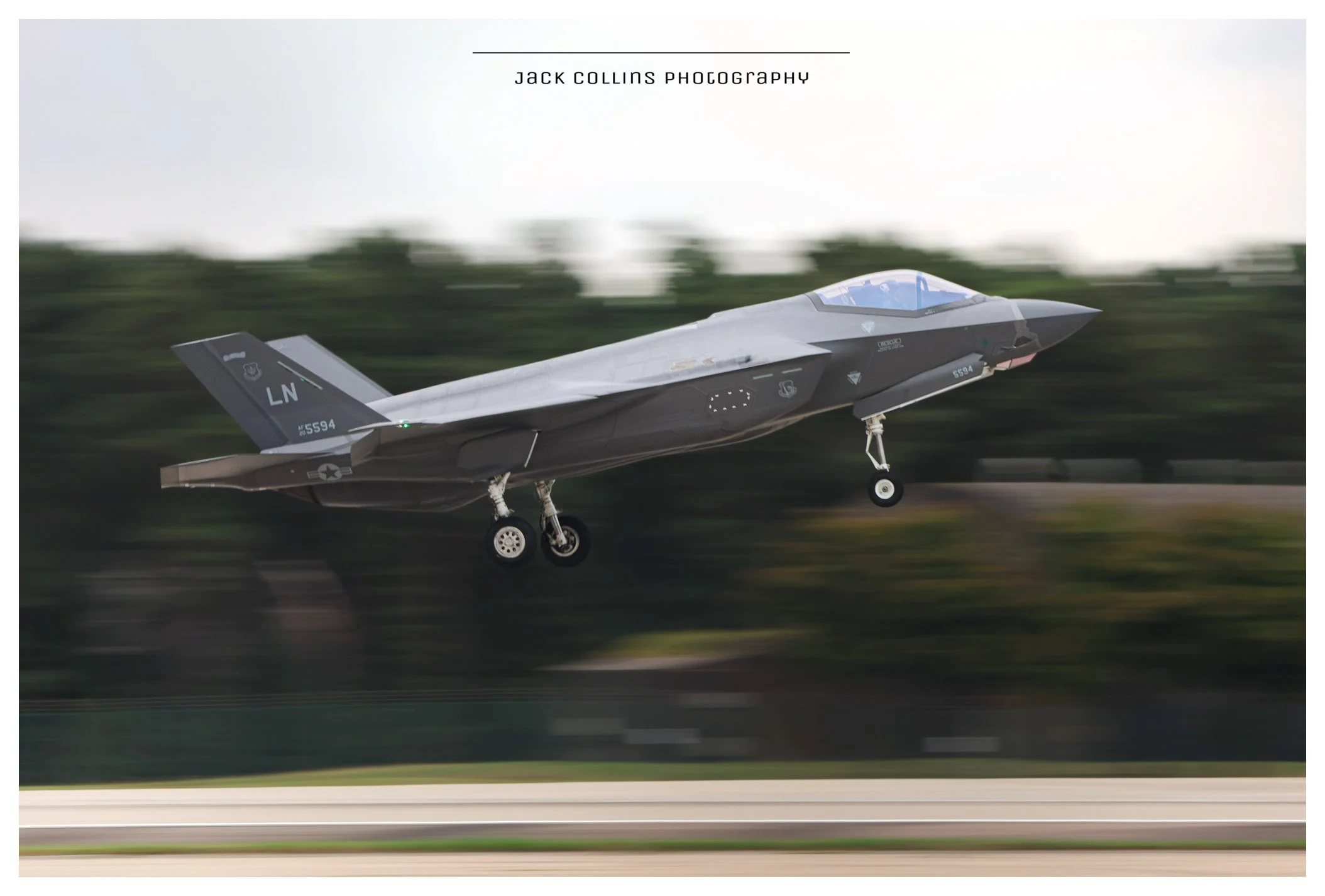 A military aircraft taking off from a runway with a blurred background of trees, featuring landing gear extended and painted in gray camouflage.