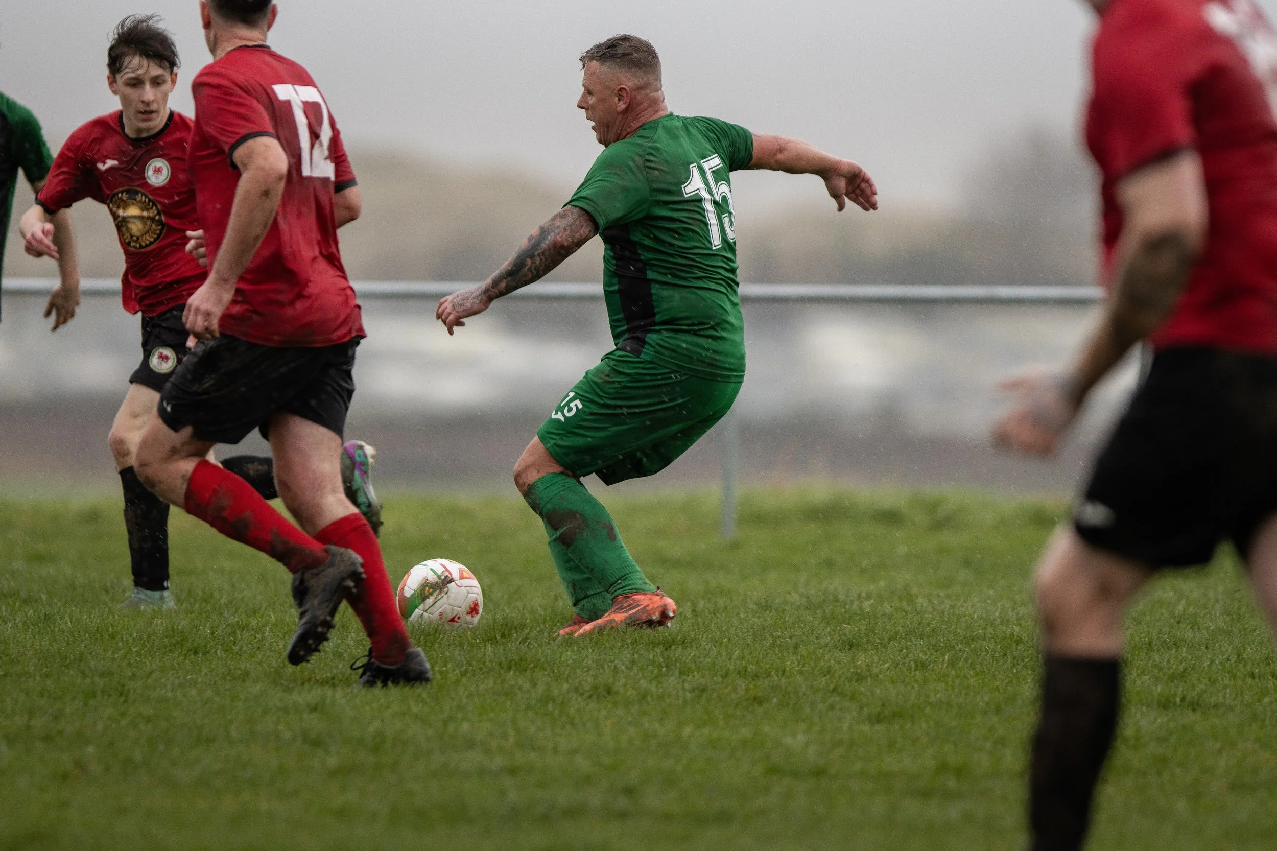 Soccer players in action on a wet field, with one in a green jersey kicking the ball while others in red jerseys defend.