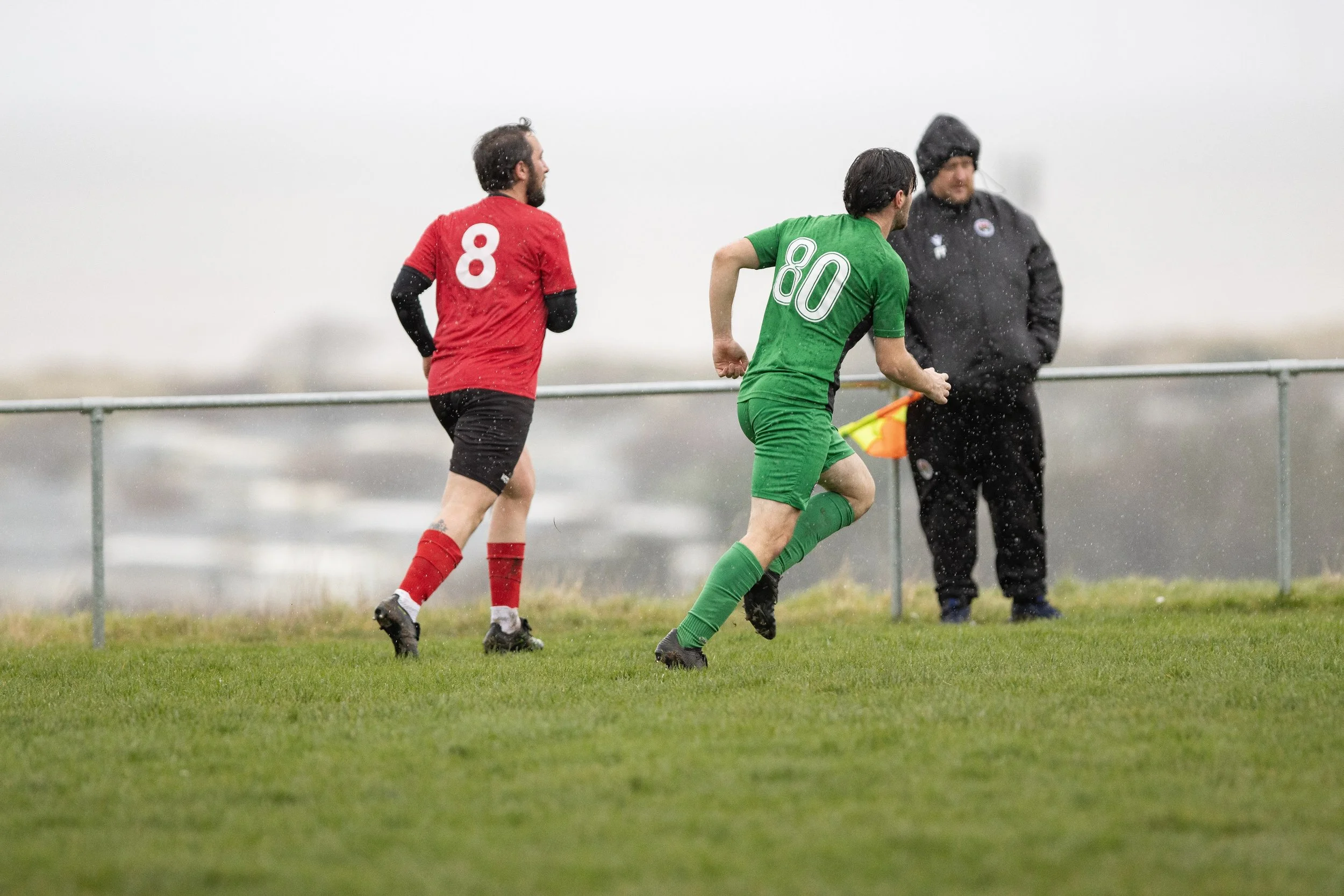 Two soccer players running on a grassy field in rainy weather, one wearing a red jersey with the number 8 and the other wearing a green jersey with the number 80, with a person in black clothing and a hat standing near the sideline.