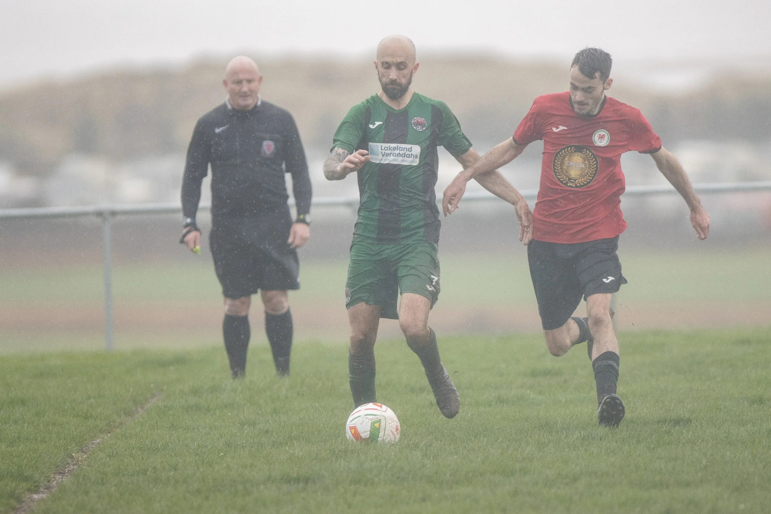 Two soccer players in green and red jerseys compete for the ball on a rainy soccer field, with a referee in the background.
