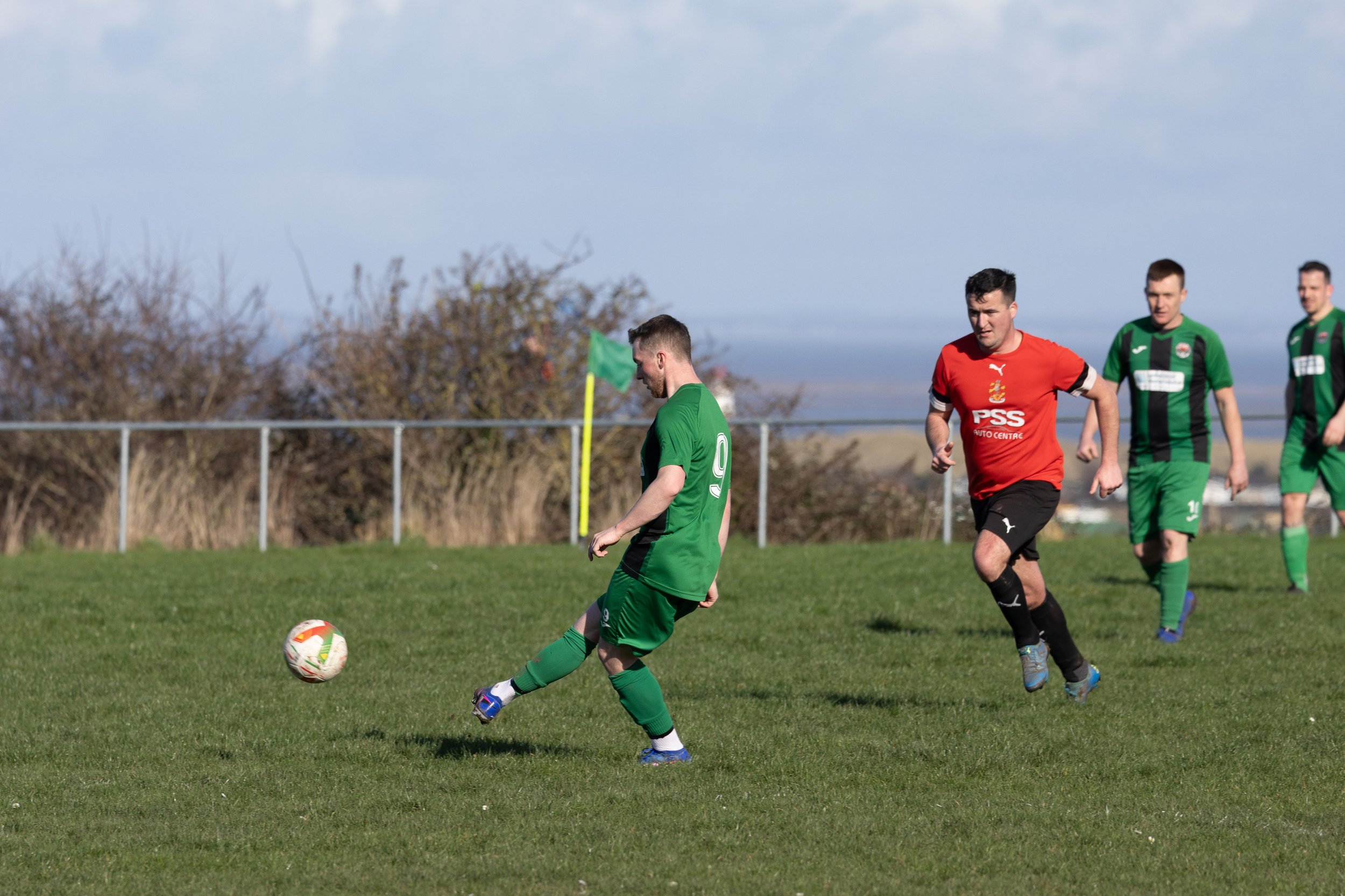 Soccer players are on a grassy field during a game, with one green team player kicking a soccer ball while three players in green and one in red are nearby, in front of a fence and bushes under a cloudy sky.