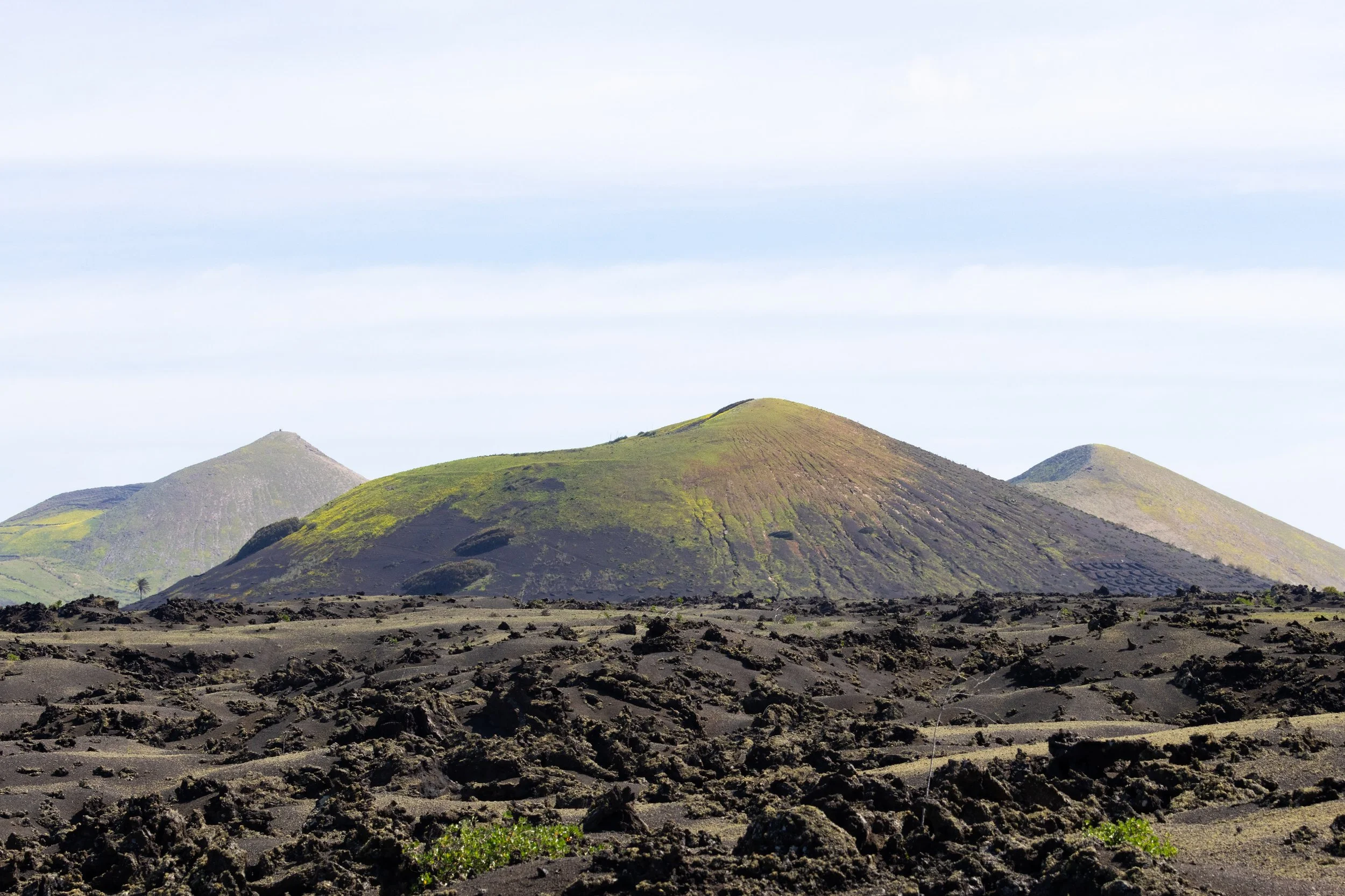 Volcanic landscape with dark, rugged rocks in the foreground and green, mossy volcanic cones in the background under a partly cloudy sky.