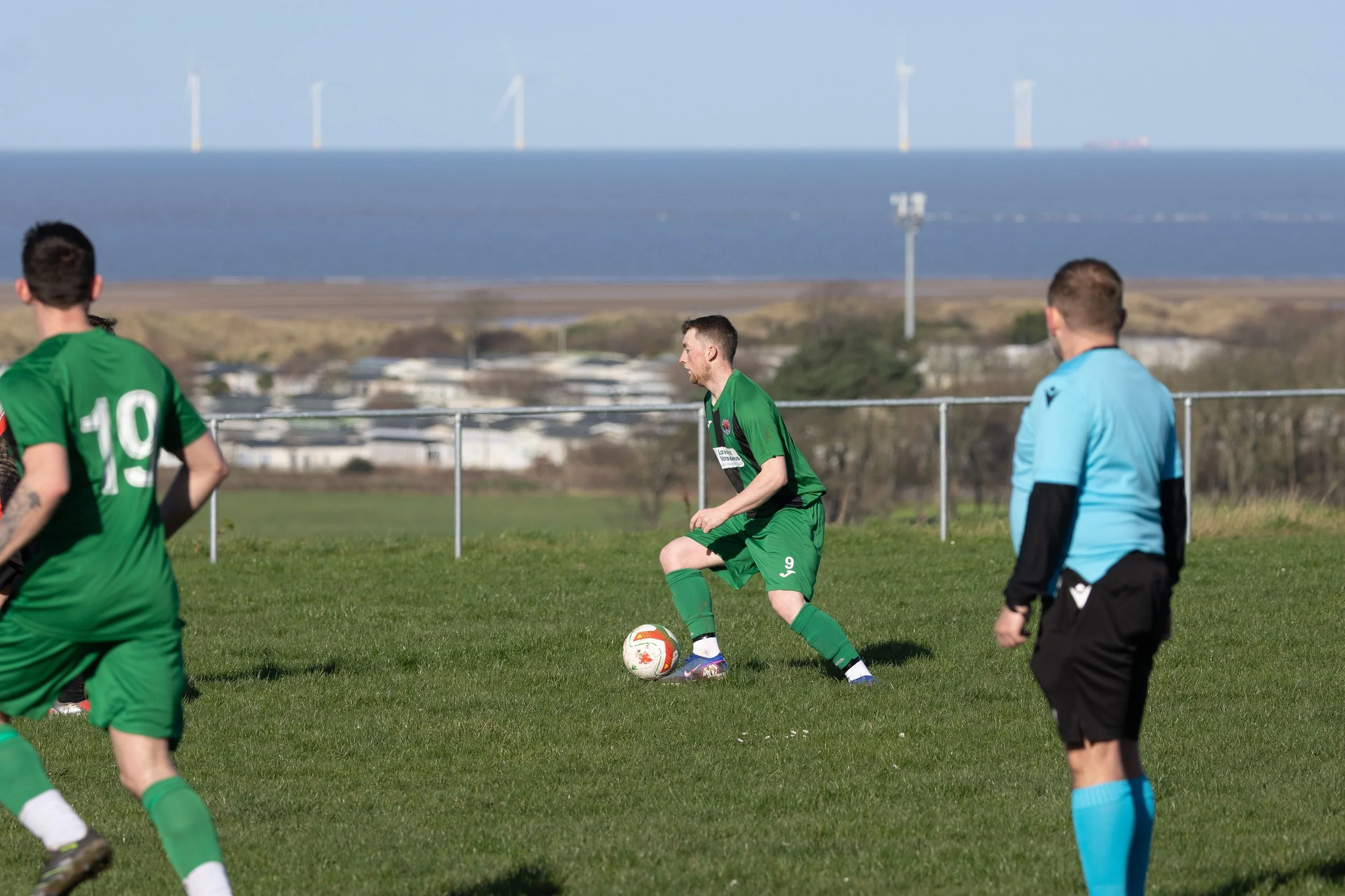 Soccer players on a green field during daytime match, with a man in a green uniform dribbling the ball near a referee in a blue uniform, background includes ocean, wind turbines, and a blue sky.