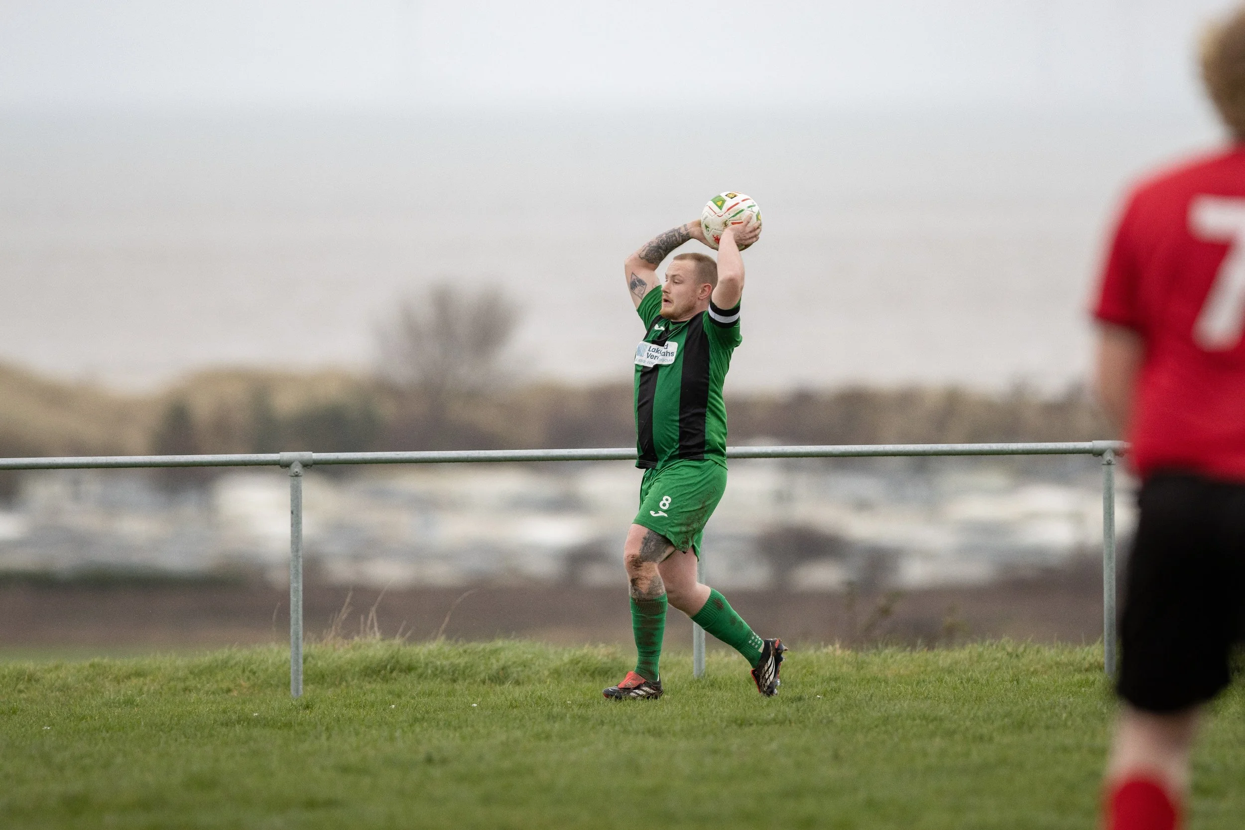 A soccer player in a green and black uniform preparing to throw the ball in during a game on a grassy field, with another player in a red jersey partially visible on the right.