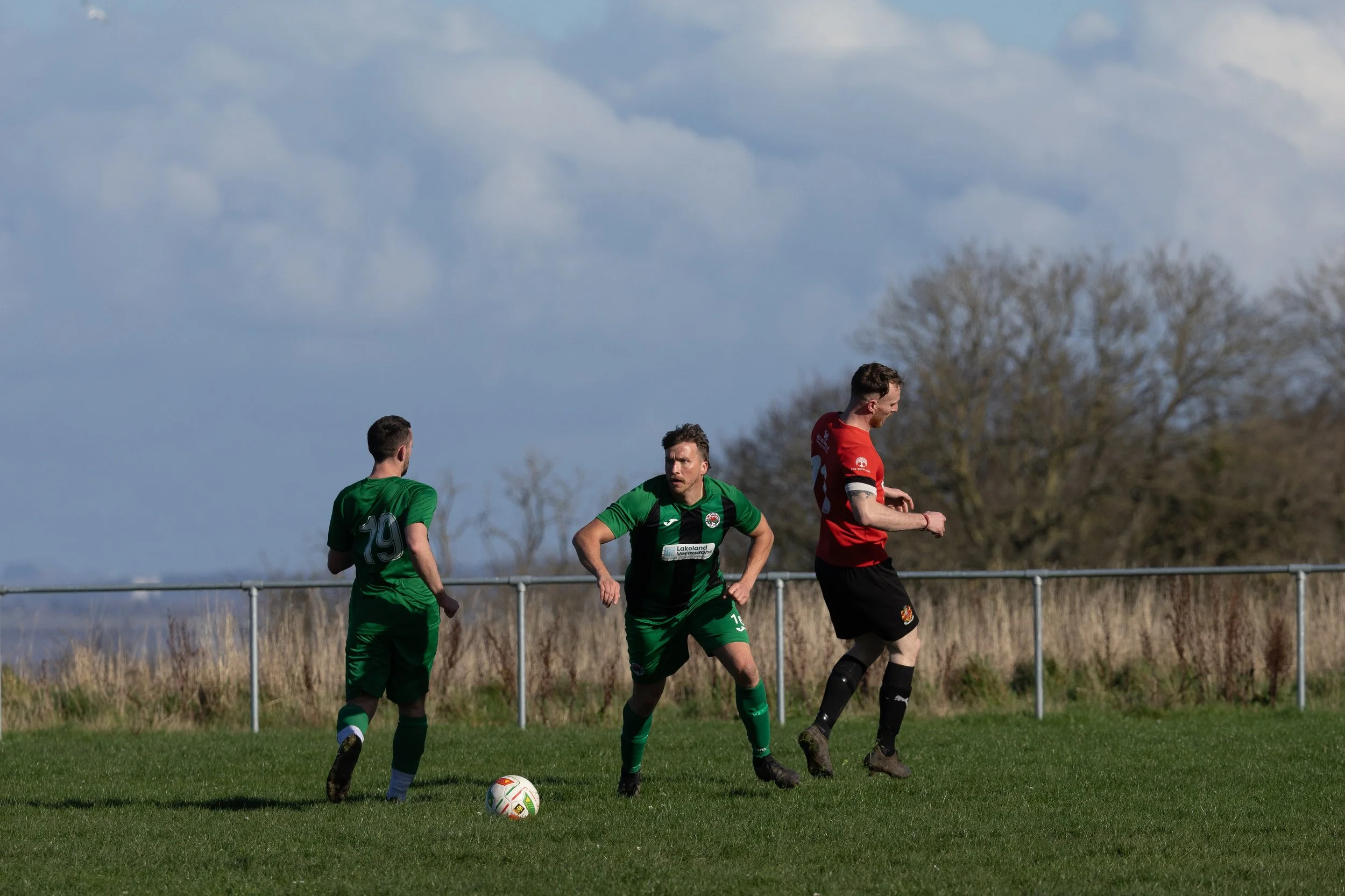 Three soccer players on the field during a match, with two in green jerseys and one in a red jersey. The player in green appears to be about to kick the ball.