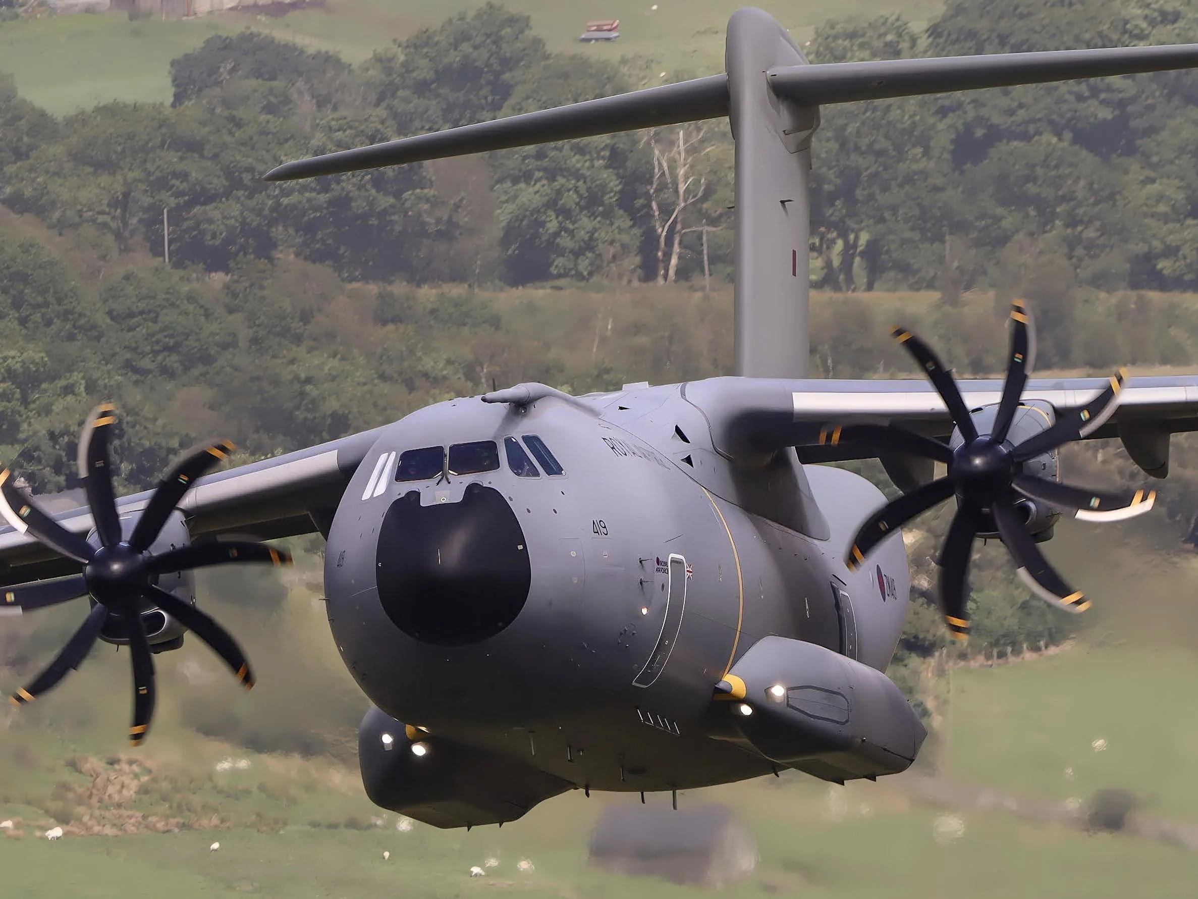 A military aircraft flying over a green landscape with trees and fields.