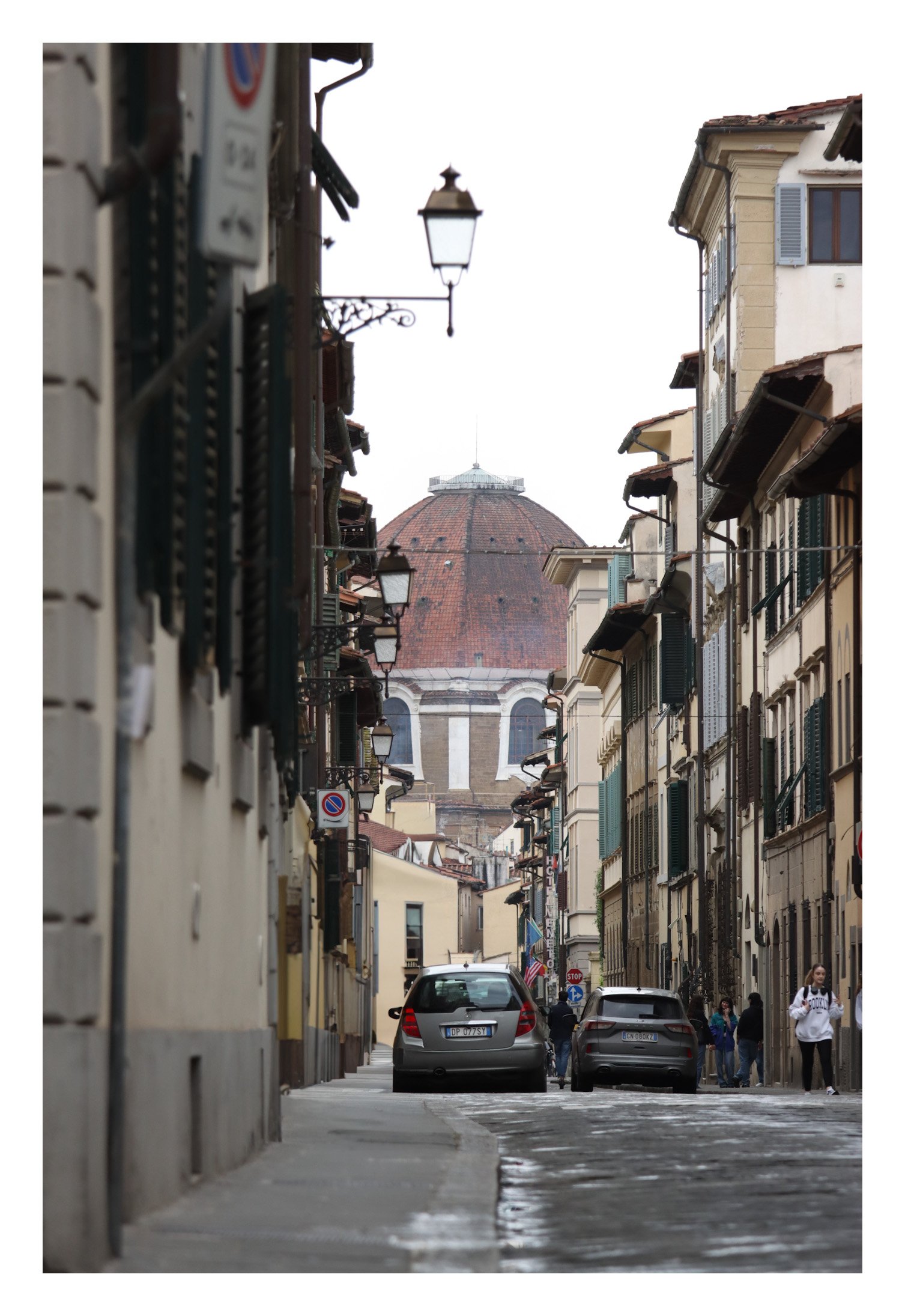 A narrow street in Florence, Italy, with parked cars, pedestrians, historic buildings with shutters, and the Florence Cathedral's dome in the background.
