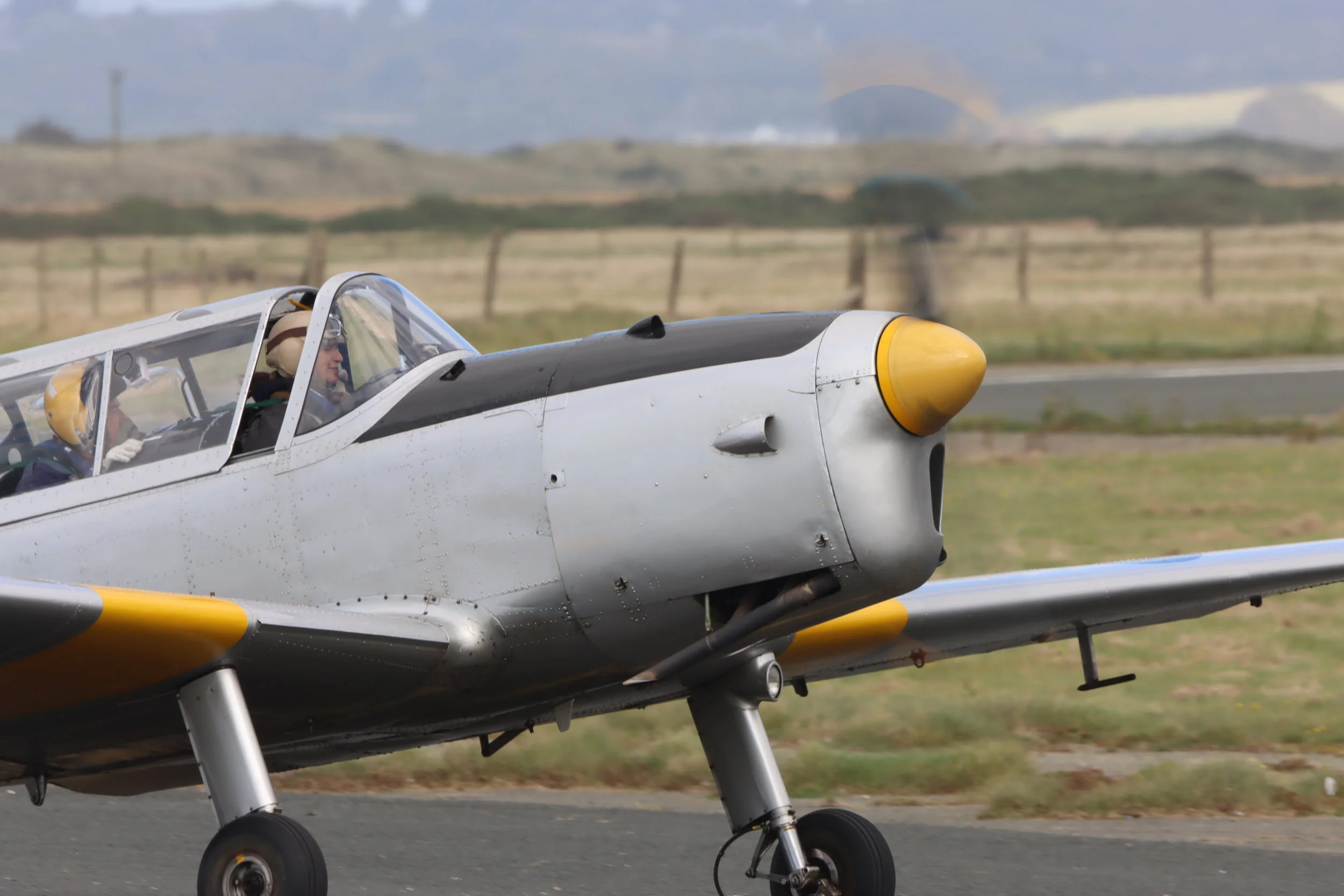 Close-up of a vintage aircraft taxiing on the runway, with pilots in the cockpit wearing helmets, and a landscape with grassy fields and mountains in the background.
