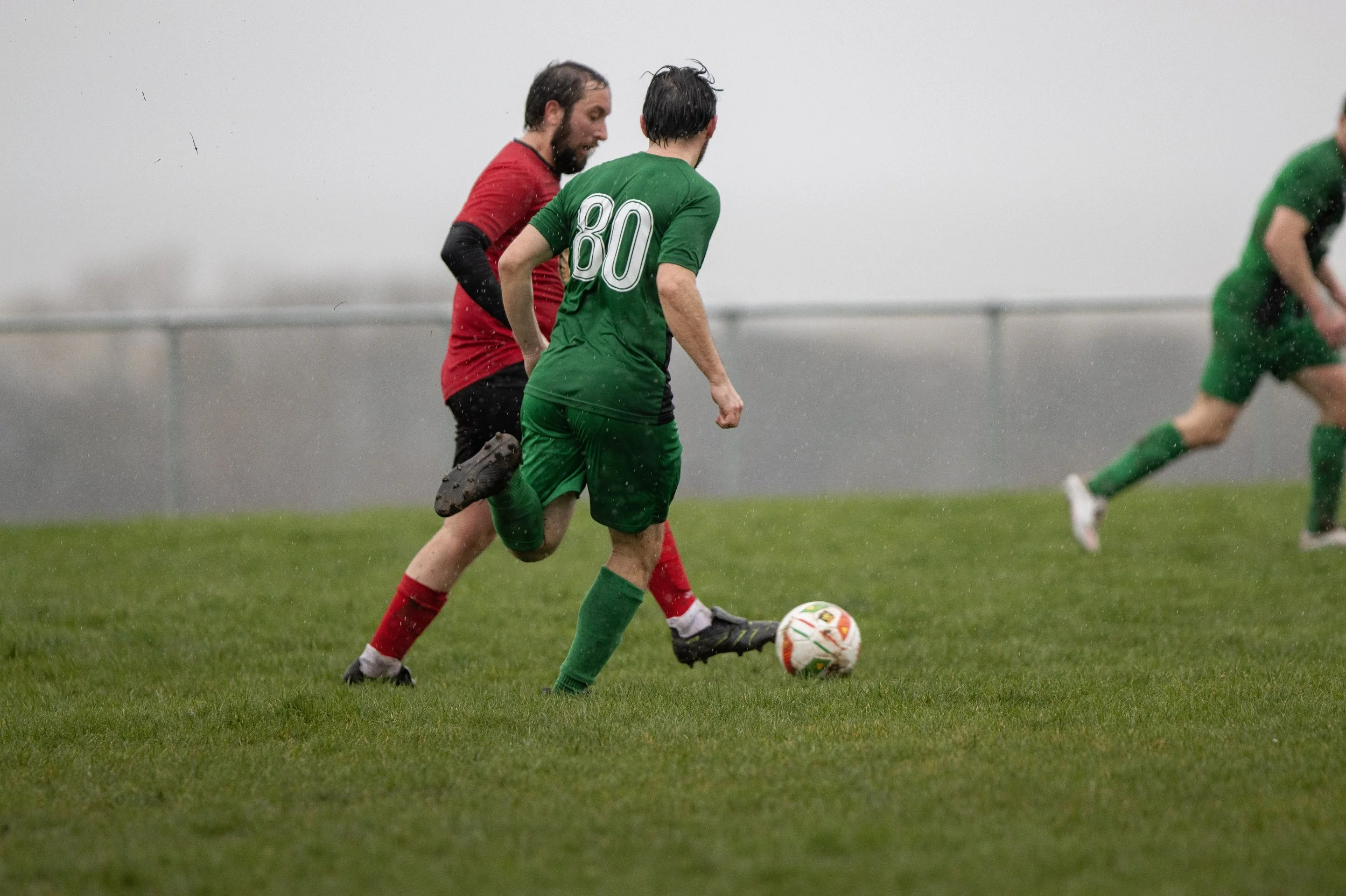Soccer players in green and red jerseys playing in the rain on a grassy field.