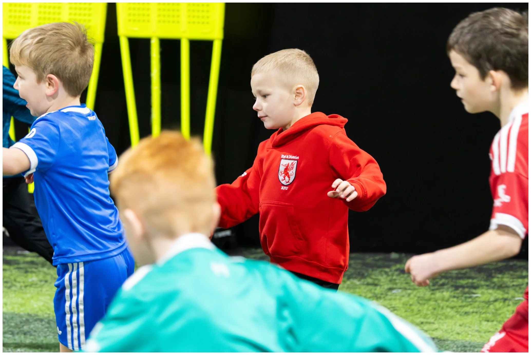 Children playing on an indoor field, some dressed in colorful sports uniforms including blue, red, and green, engaging in a game or activity.