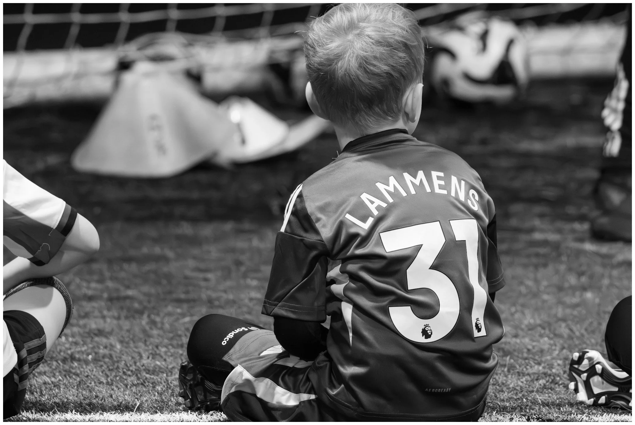 A young boy sitting on the grass during a soccer game, wearing a jersey with the name 'Lammens' and the number '31' on the back, with soccer cleats and a soccer ball nearby.