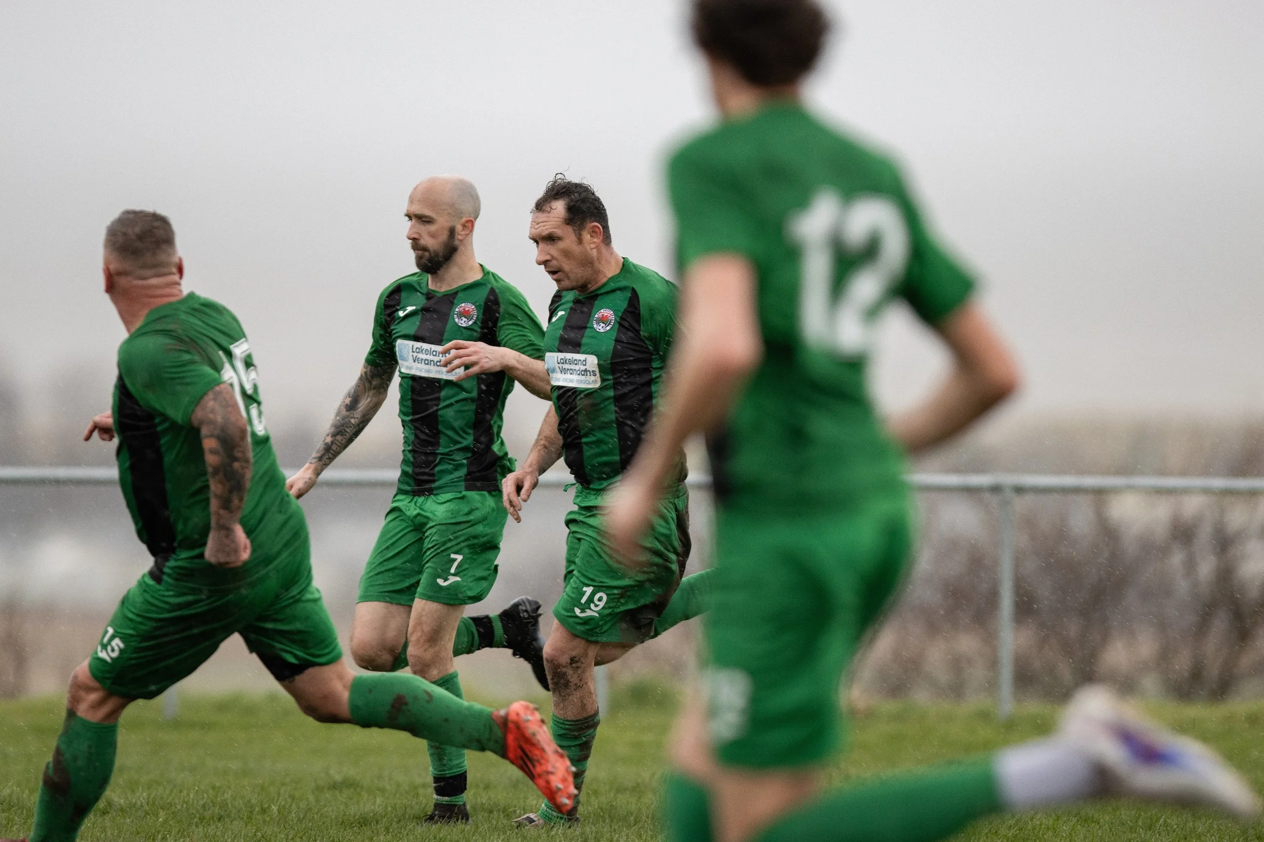 A soccer match with players in green jerseys on a muddy field in rainy weather, with one player in focus and the others slightly blurred.