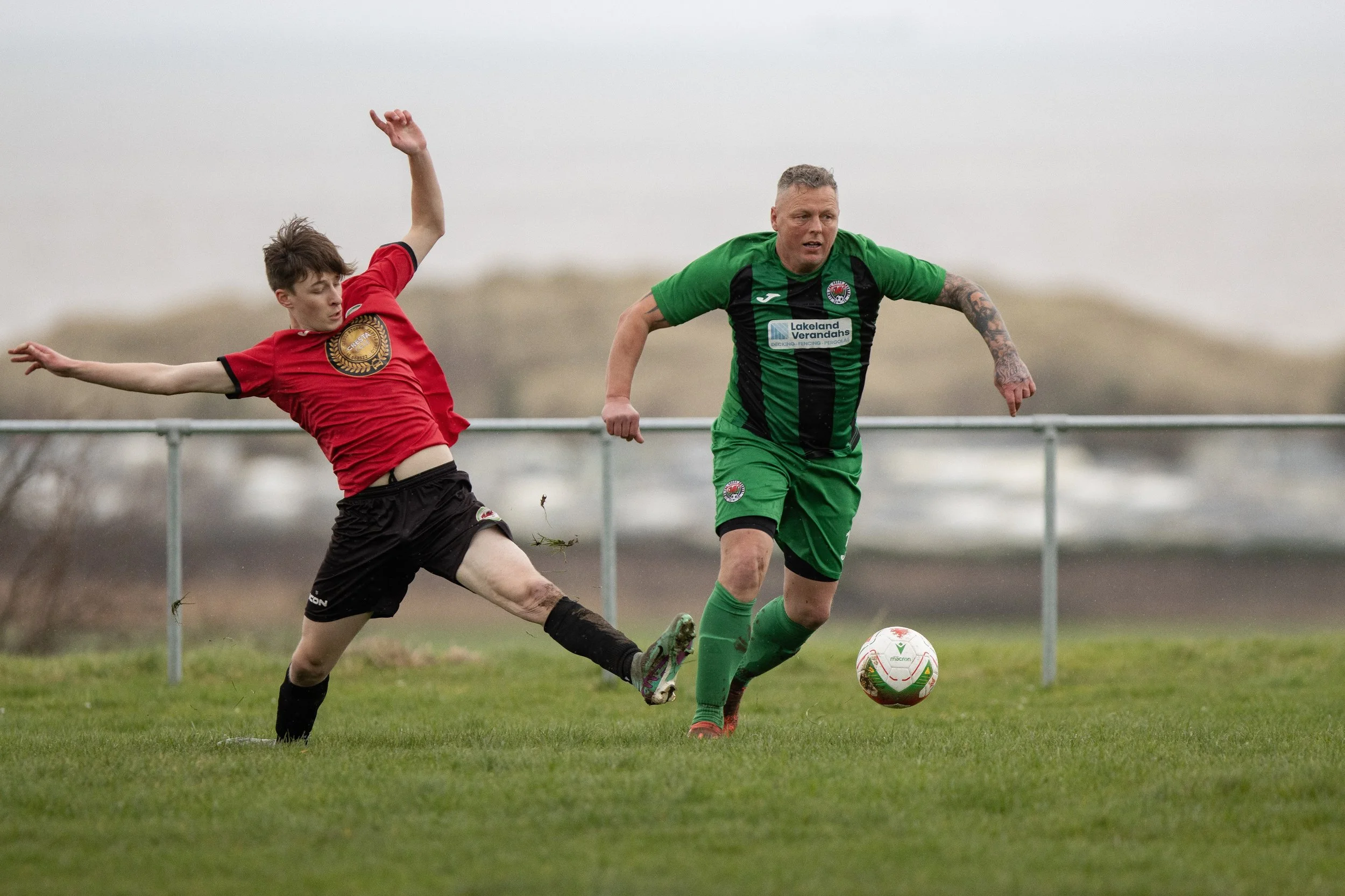 Two soccer players in action on a grassy field, one in a red and black uniform attempting to kick the ball while the other in a green and black uniform runs after it, with a cloudy sky in the background.
