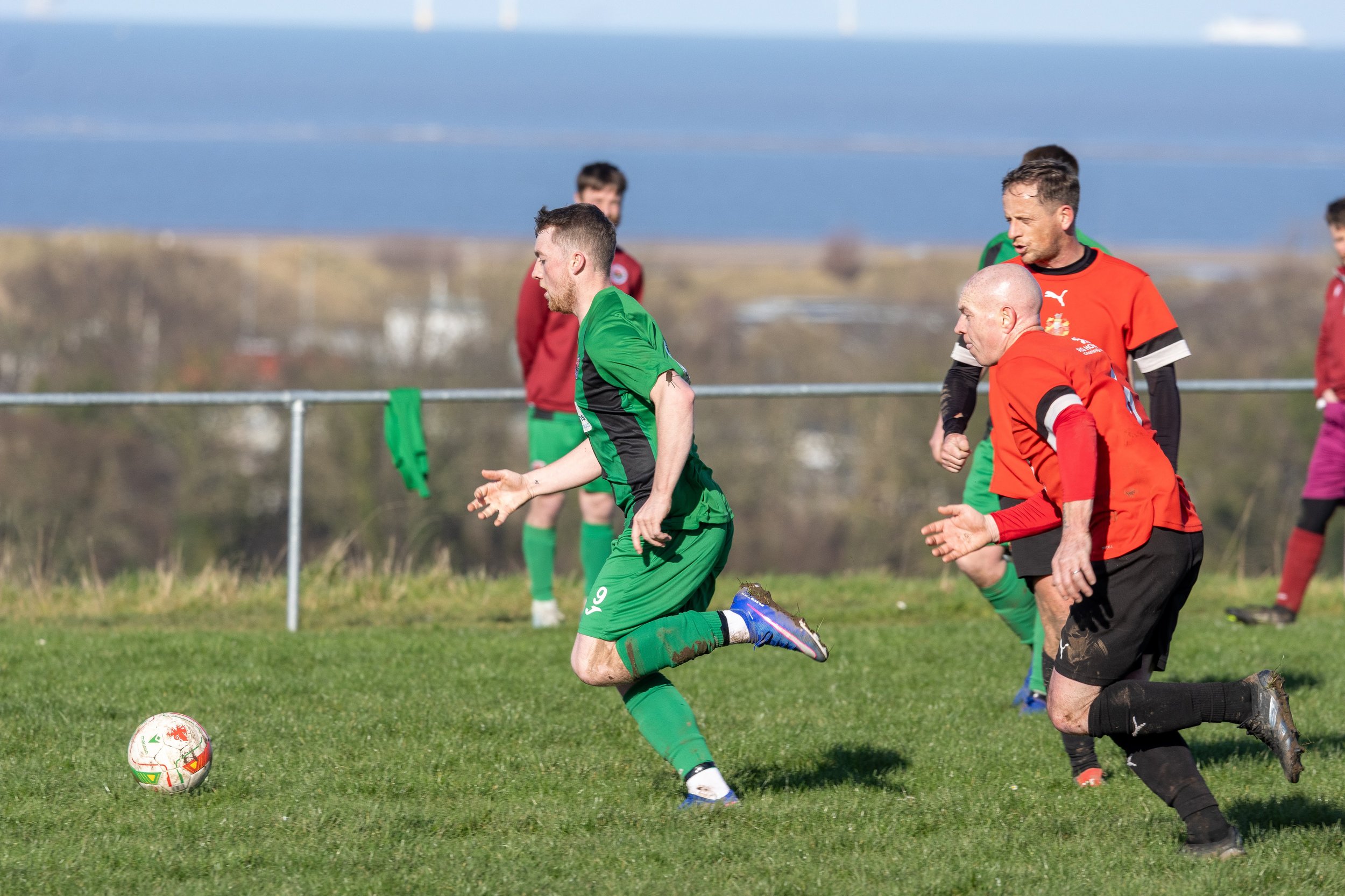 Soccer players in green and red uniforms competing on the field, with a soccer ball in the foreground and a clear sky in the background.