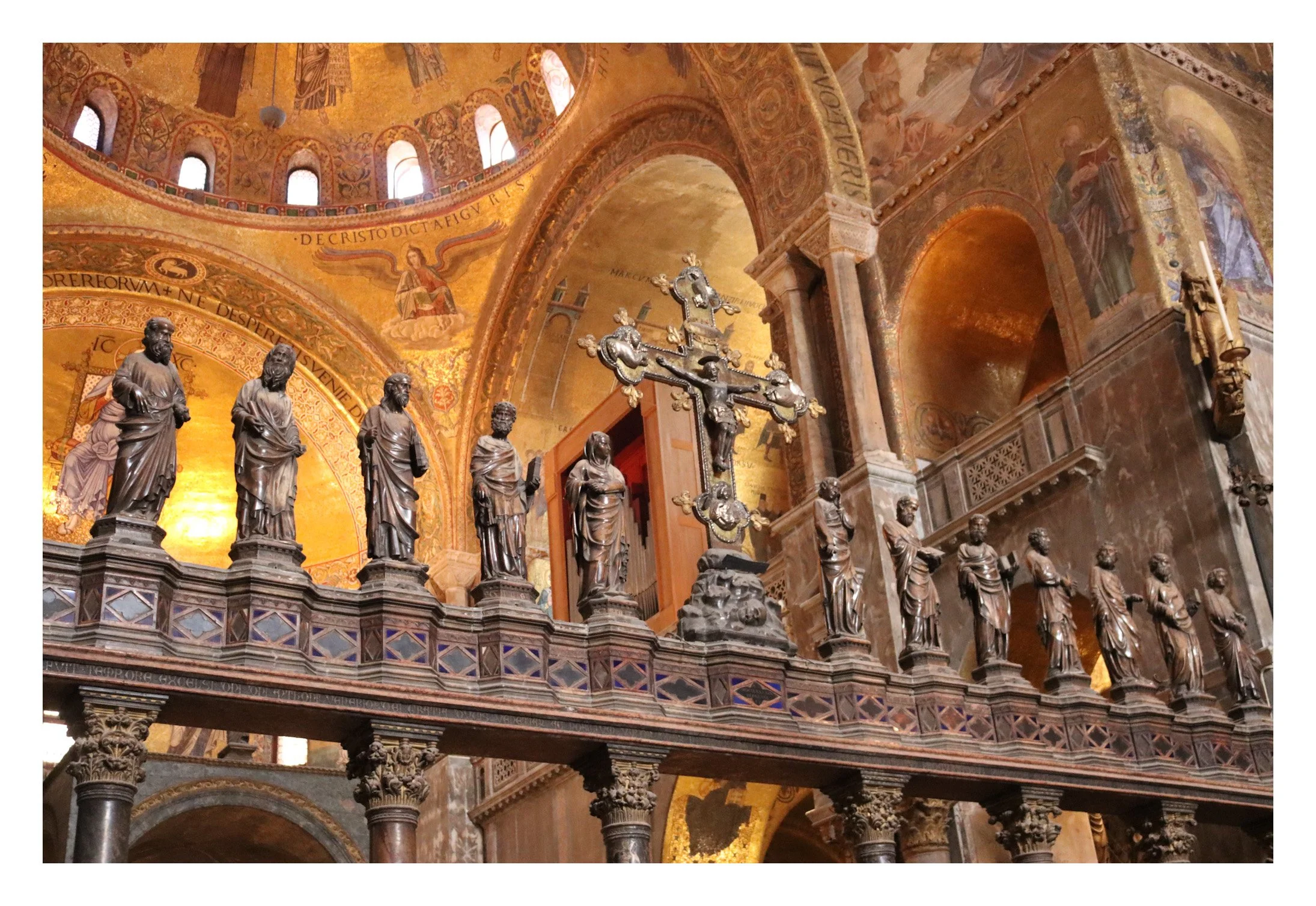 Inside a church with a gold mosaic ceiling, a crucifix and a series of statues of saints on a decorative balcony.