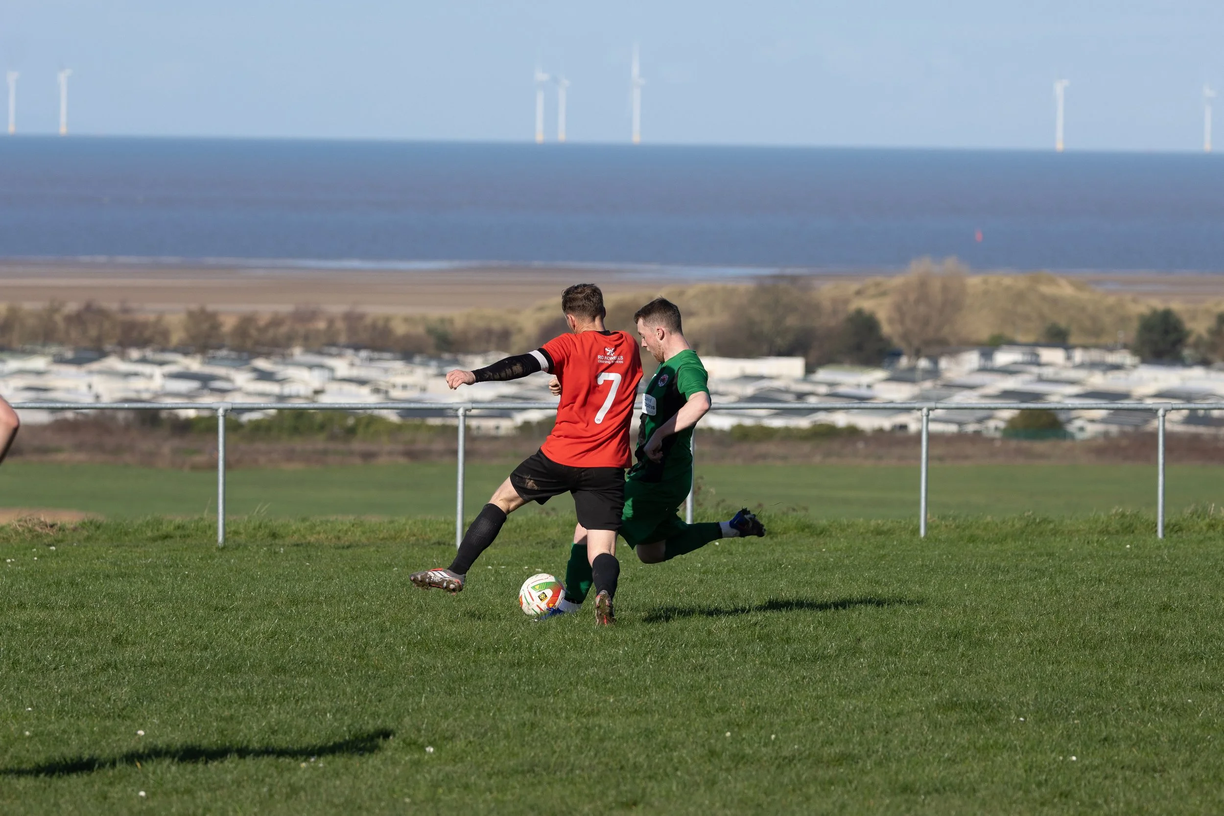 Two soccer players, one in red and one in green, competing for the ball on a grassy field with a rural landscape and wind turbines in the background.