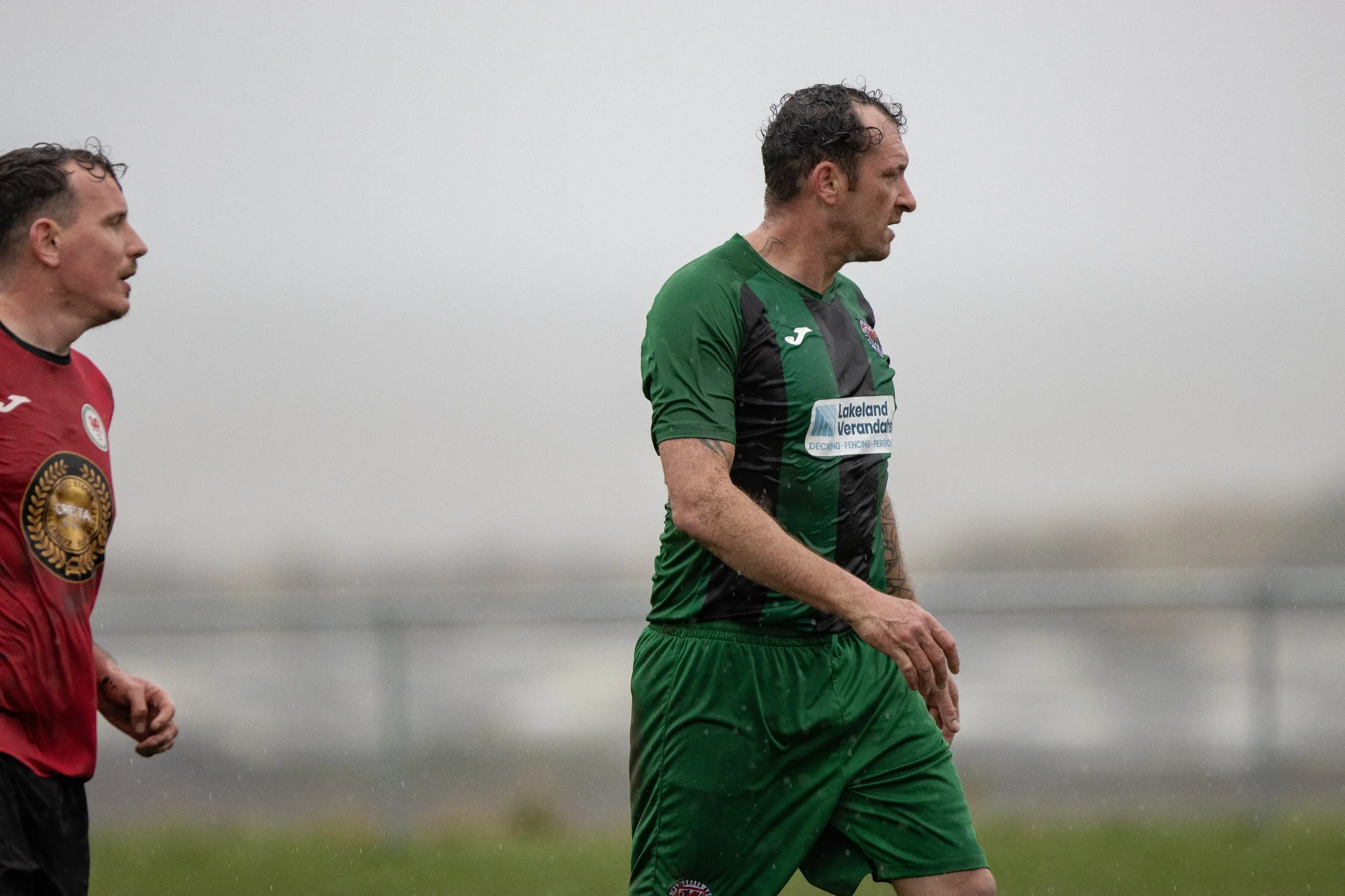 Two soccer players walking on a rainy field with a gray sky, one in a red jersey and the other in a green and black jersey.