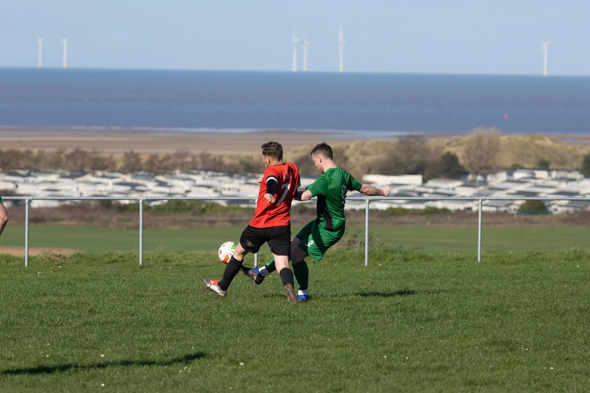 Two men playing soccer outdoors on a grassy field, with a body of water and wind turbines in the background.