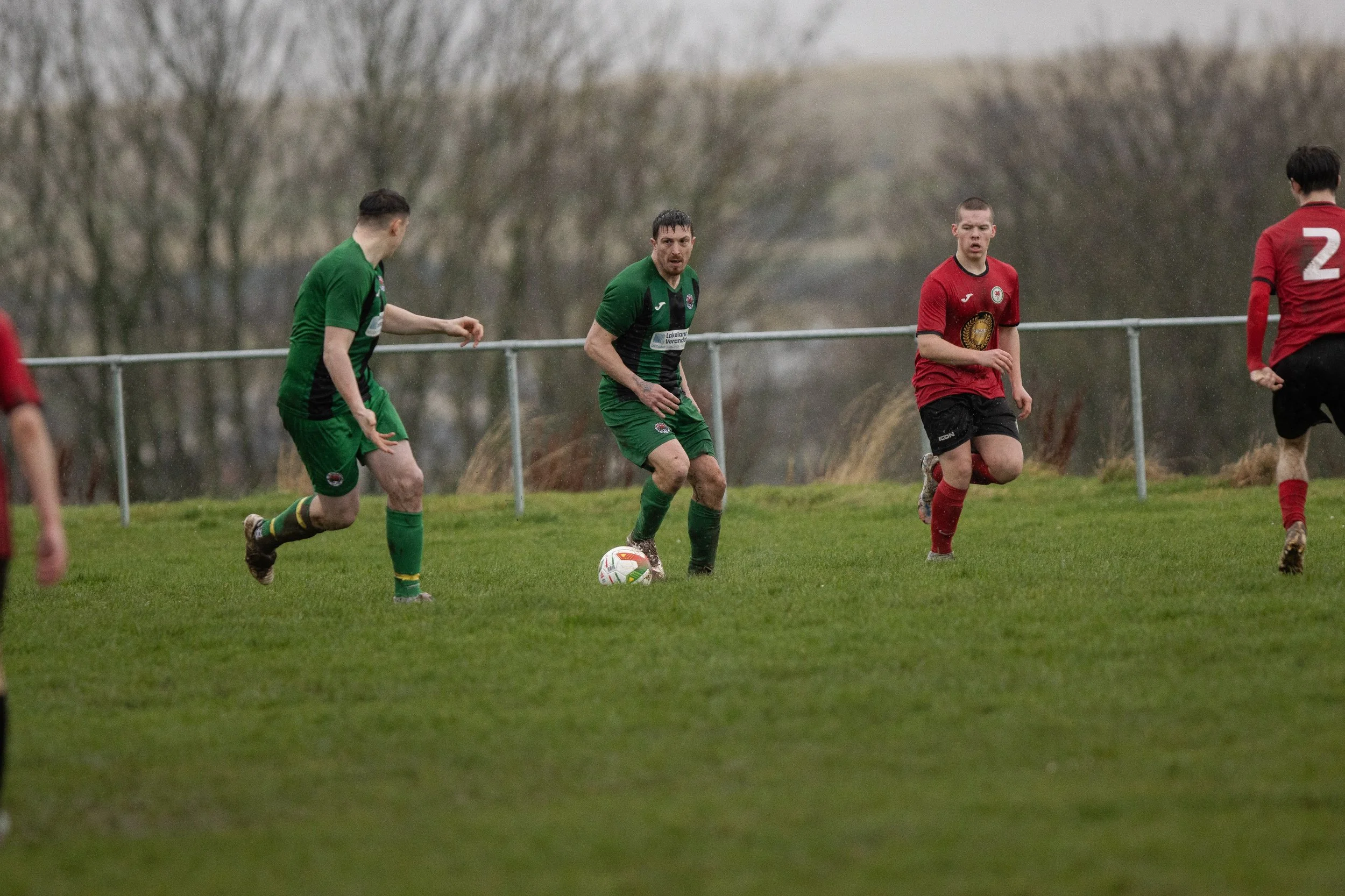 Soccer players on a grassy field, with the player in green in the center preparing to kick the ball, and other players in red nearby, during an overcast day.
