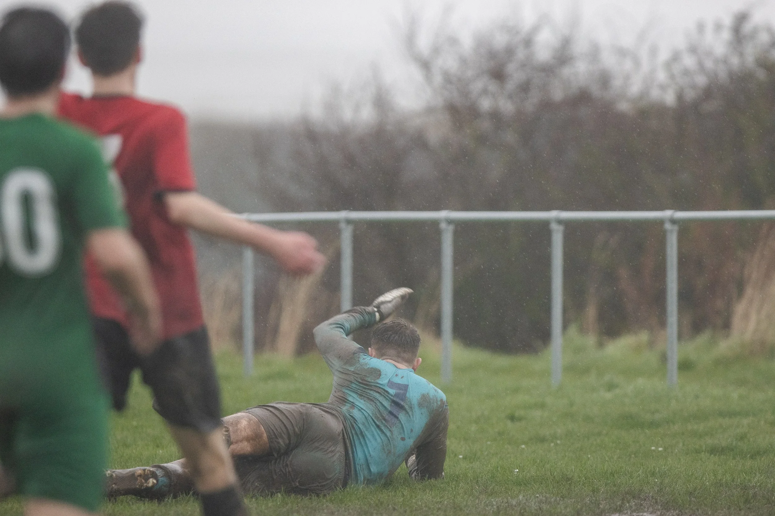 A soccer game in rain with a goalkeeper diving on the ground, wearing a muddy blue jersey, while two outfield players, one in green and one in red, watch nearby.