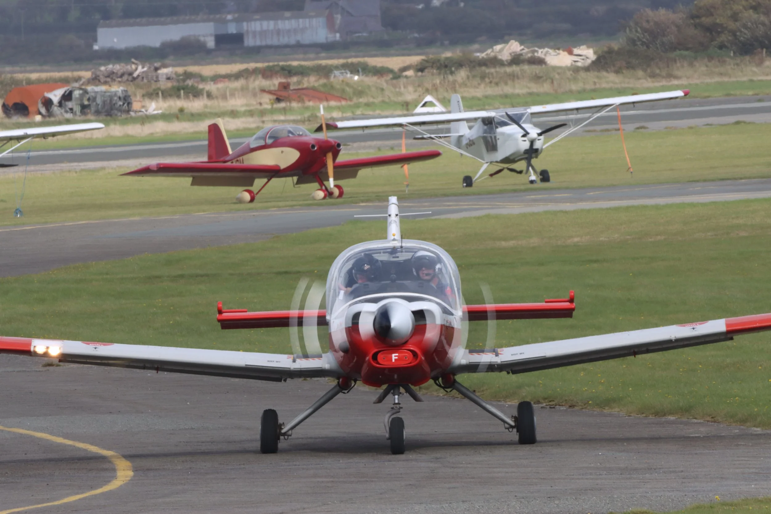 Small display aircraft on runway with two small planes in background, one red and one white, and aircraft wreckage and grass in distance.