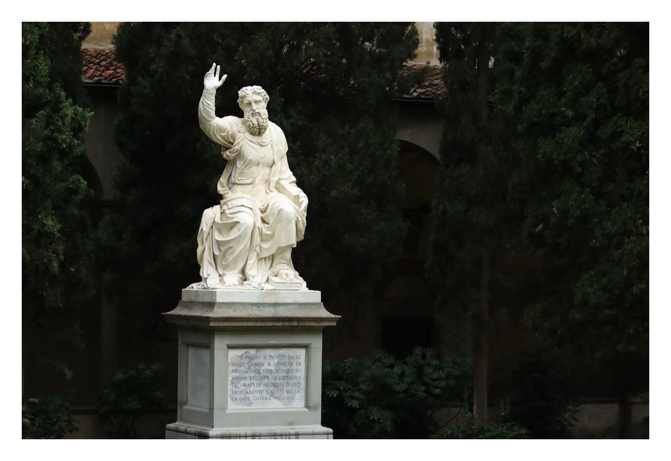 A white marble statue of a seated bearded man with curly hair, wearing robes, raising his right hand in a gesture of greeting or blessing, set outdoors amidst greenery with trees and a building in the background.