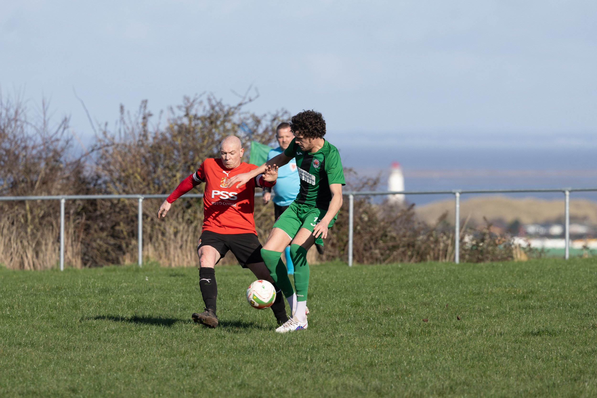 Two soccer players competing for the ball on a grassy field, with a referee and trees in the background, and a lighthouse on the horizon.